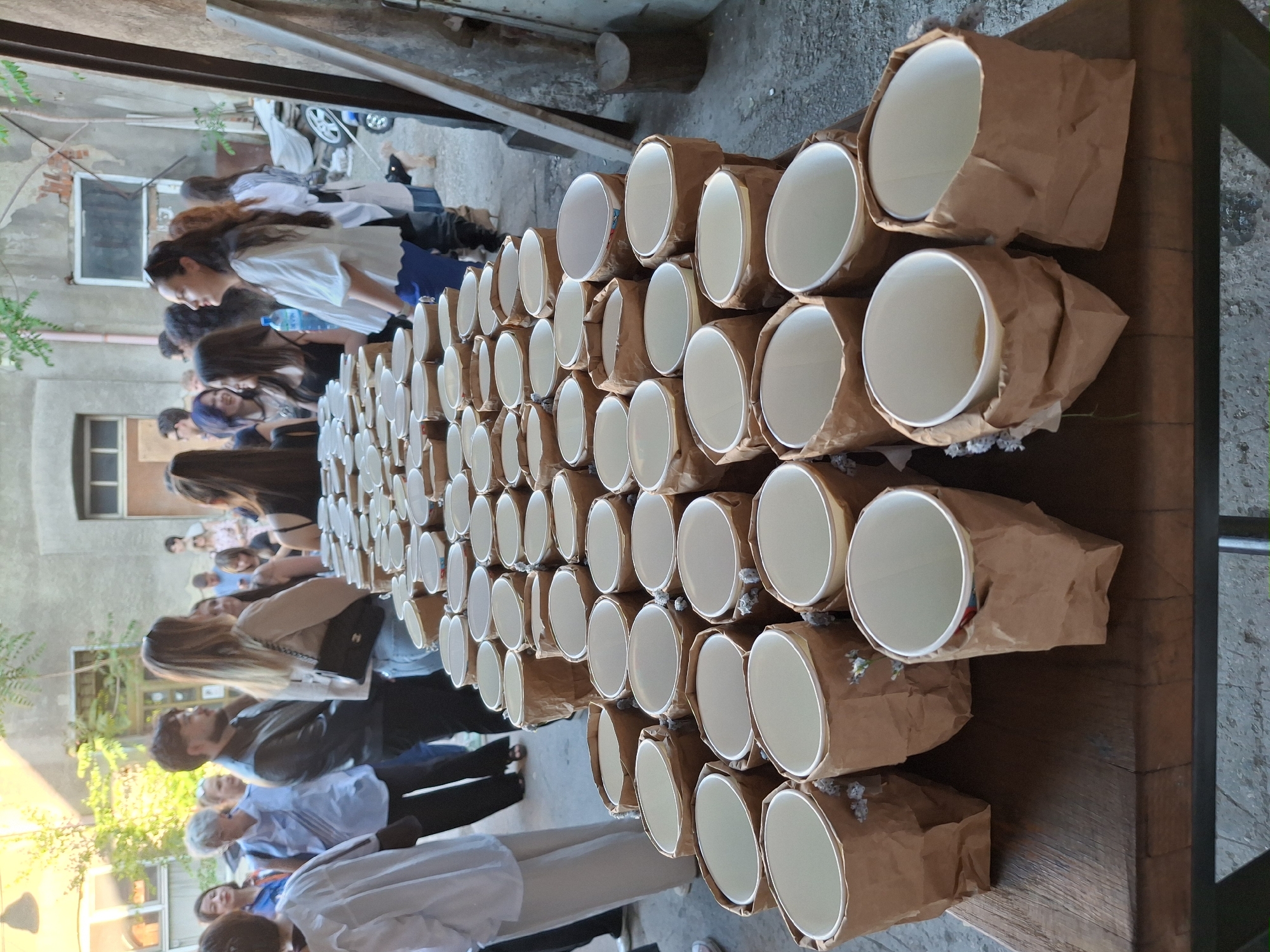 The image shows a table laden with many paper cups, each encased in a brown paper bag, likely for some beverage distribution. A line of people stands beside the table, apparently waiting to receive the beverages. The scene appears to be an outdoor gathering within a city environment, as suggested by the surrounding buildings and natural light, but the specific city is unknown.