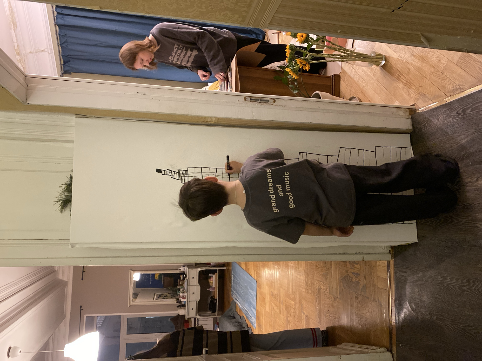 A medium shot shows a boy drawing on a large white surface with a black marker. He is wearing a grey sweatshirt with the words "grand dreams and good music" printed on it, along with black pants. He stands in a doorway, and the drawing resembles a cityscape with numerous rectangular shapes.
Through the doorway, in the background, a person with reddish-brown hair is leaning over a desk or table, appearing to be organizing or sorting items, with a vase of yellow sunflowers nearby. The room beyond the doorway has hardwood floors.
Through another doorway in the background, an interior with a lighter color scheme can be seen. There are wooden floors, furniture, and what appears to be a mirror or a framed picture on the wall.
The location appears to be an indoor space, likely a private residence or apartment. There are no visual cues or specific details in the image to confirm the city of Moscow or its location in Russia.