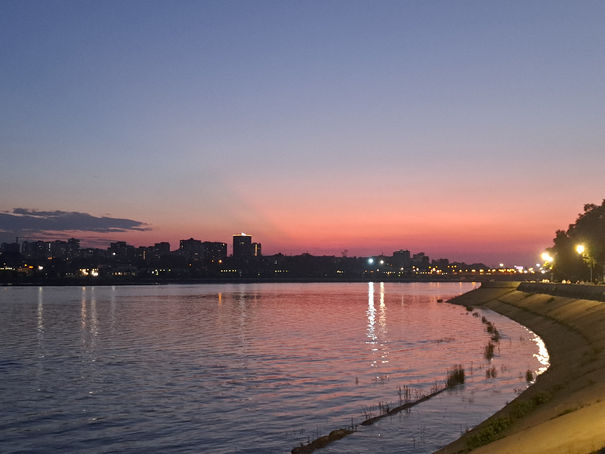 This is a twilight photograph of the Angara River in Irkutsk, Russia. The sky displays a gradient of colors, transitioning from a pale blue at the top to a vibrant pink and orange near the horizon. Silhouetted against the colorful sky is the city skyline, characterized by numerous buildings and the illuminated windows of some structures. The river's surface reflects the sky's colors and the city lights, creating shimmering patterns on the water. The right side of the image features a paved embankment with lampposts casting warm light. There are no visible people in the foreground or midground.