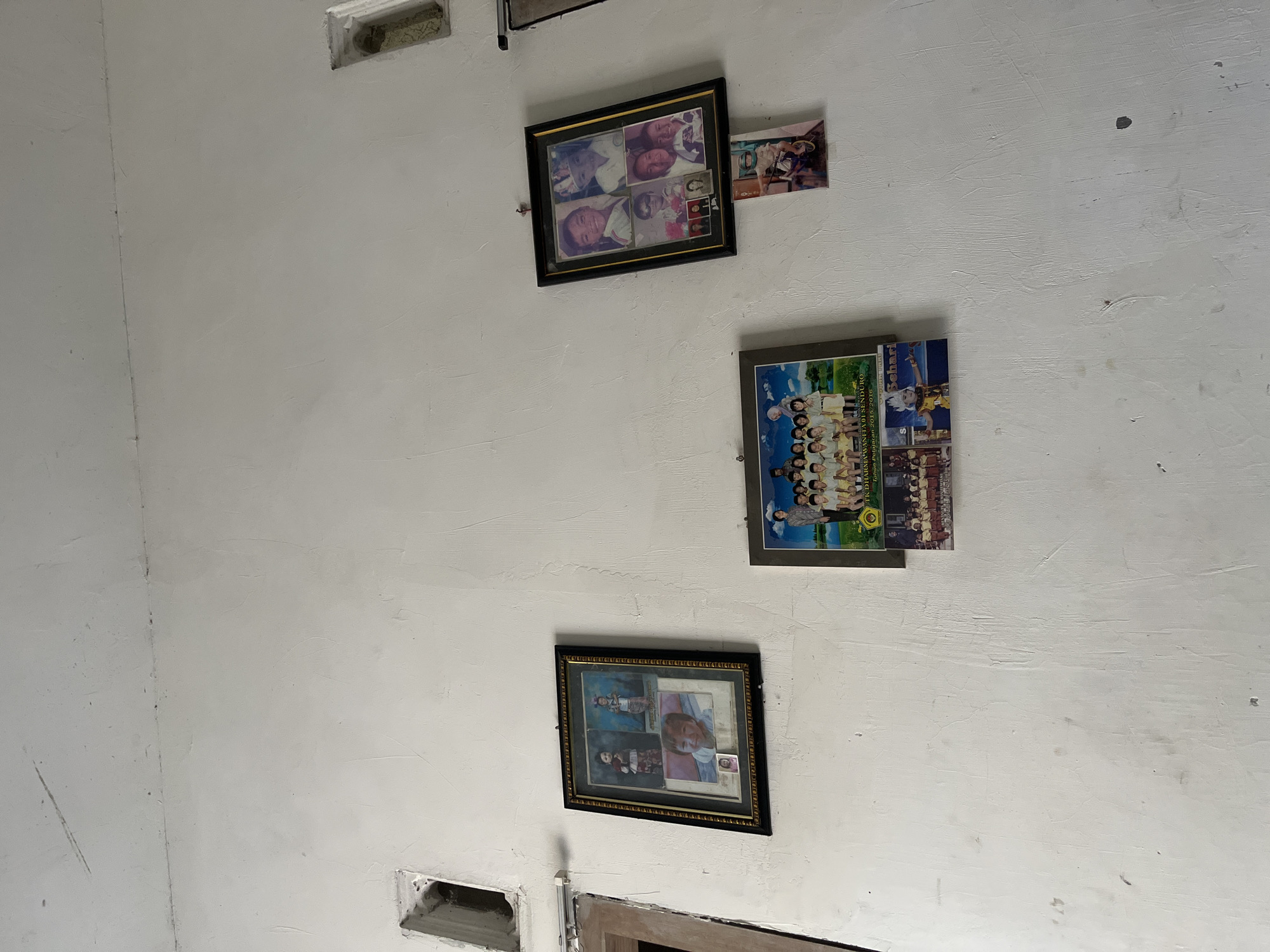 A white, textured wall in Pasrukkrajan Satu, Indonesia, displays several framed photographs and a small, ajar window opening near the top left of the image. To the left of the center, a black-framed collage of four portraits shows individuals in formal attire. Below this collage is a smaller framed photograph of a group of children in uniform. To the right, a black-framed collage of five portraits features children wearing a variety of outfits. Below this, a single, small rectangular photograph depicts a child on a motorbike. Further to the right and slightly below the portrait collage, a small, squared framed photo shows three individuals. The wall itself appears somewhat aged, with subtle imperfections and shadows contributing to its textured appearance.