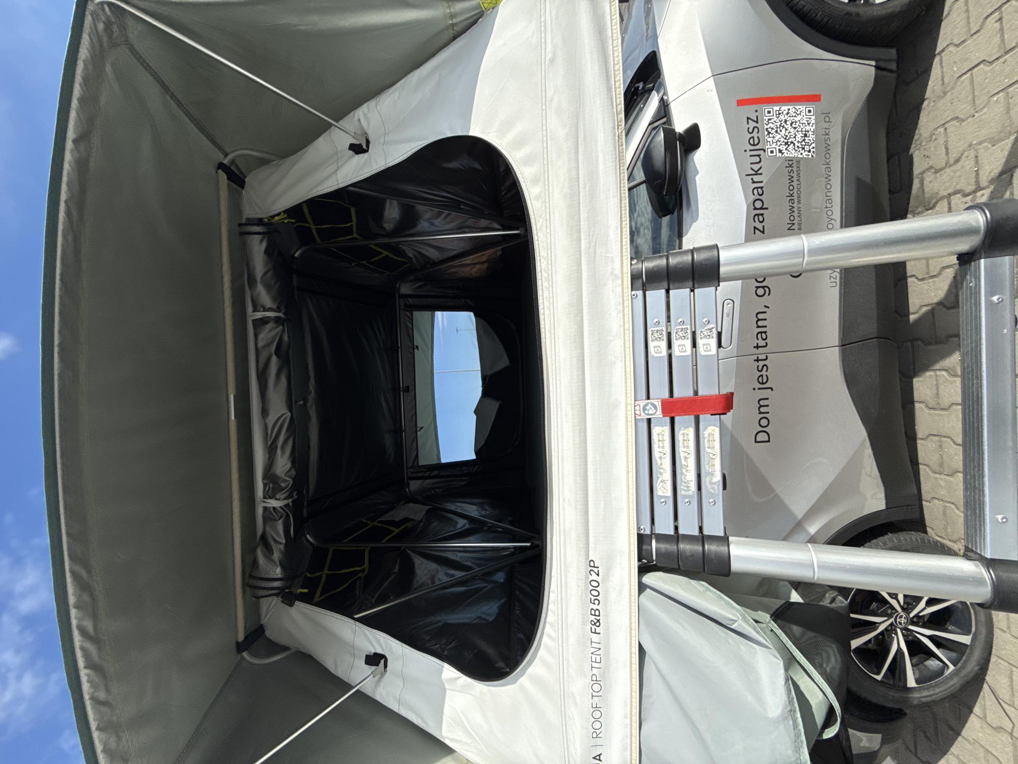 The image shows a rooftop tent set up on a silver car. The tent is open, revealing the interior. A ladder is attached to the side of the car, leading up to the tent. The car is parked on a paved surface. On the side of the car, there is text that reads "Dom jest tam, gdzie zaparkujesz. Nowakoswski BIELANY WROCŁAWSKIE uzy.oyotanowakowski.pl" and a QR code. The text indicates the location is in Wrocław, Poland. The sky is blue with some clouds.