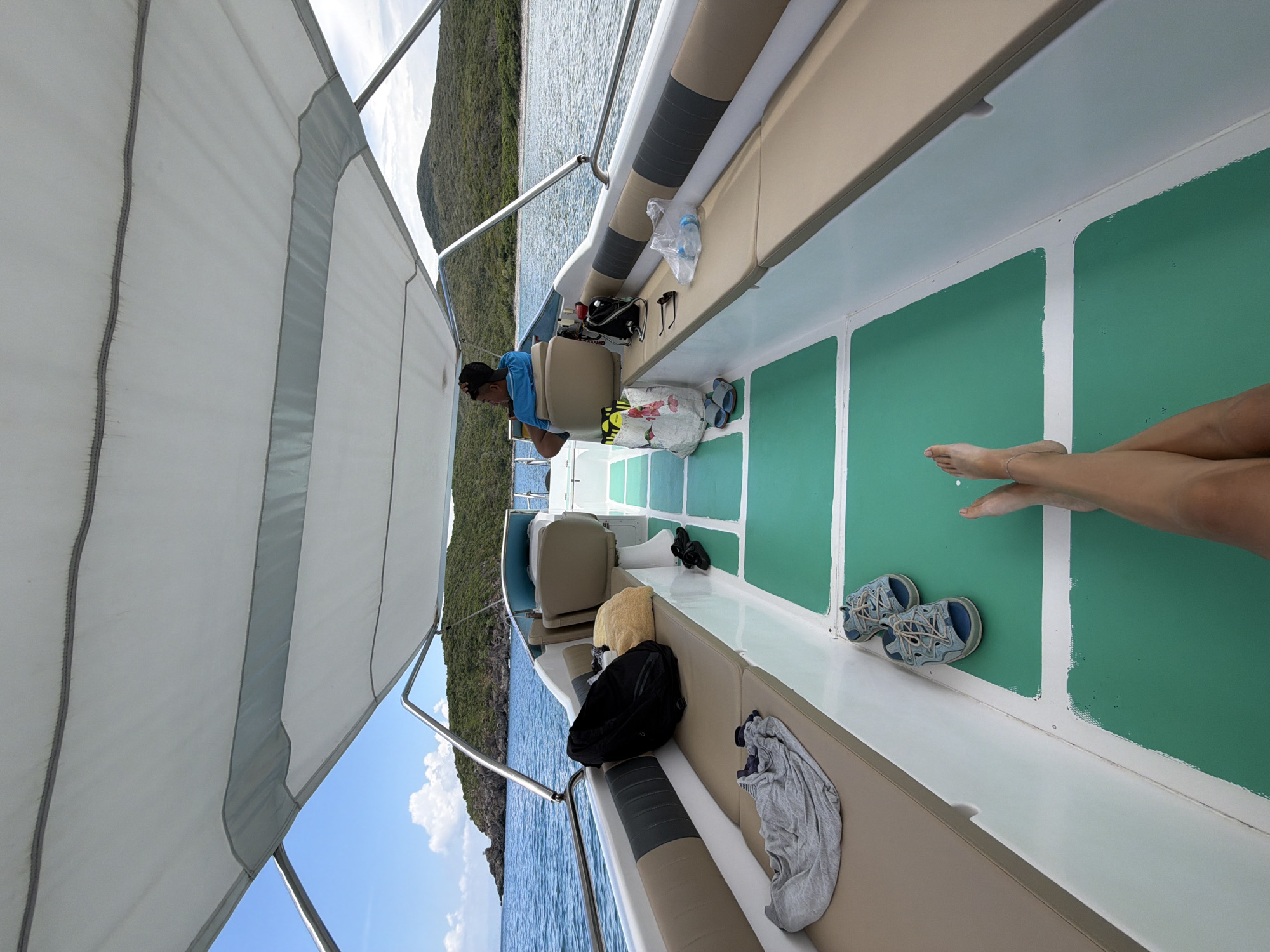 The image depicts the interior of a boat, possibly near Bac Nha Trang Ward, Vietnam, as suggested by the coastal scenery visible. A man in a blue shirt sits at the helm, focused on something out of view. Next to him are water bottles and other personal items. On the opposite side, bare legs extend into the frame. A pair of shoes are on the floor.