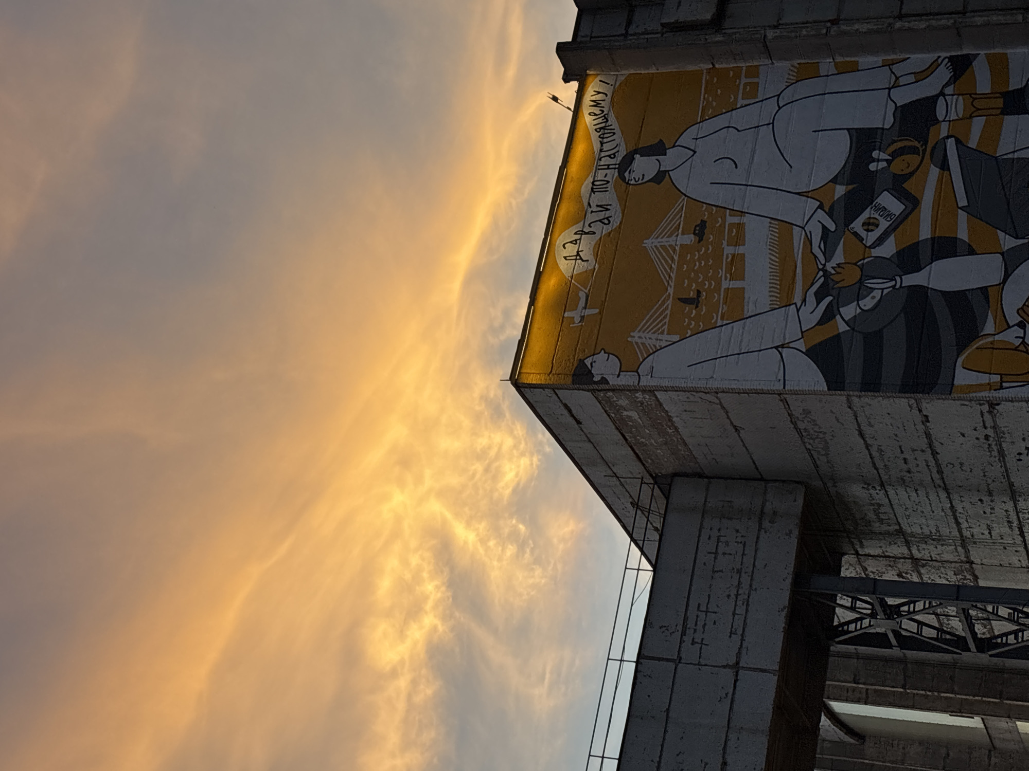 The image shows a mural on the side of a concrete building at dusk in Saint Petersburg, Russia. The mural depicts two figures, a man and a woman, sitting with their hands touching. They are shown against a backdrop of a bridge, boats, and birds. Above them, the sky is illuminated with a vibrant orange and yellow sunset, with wispy clouds stretching across the frame. The building itself is weathered concrete, and a metal walkway or bridge is visible on the left side of the image, extending from the building. The overall scene is one of urban decay contrasted with the beauty of the natural sky.