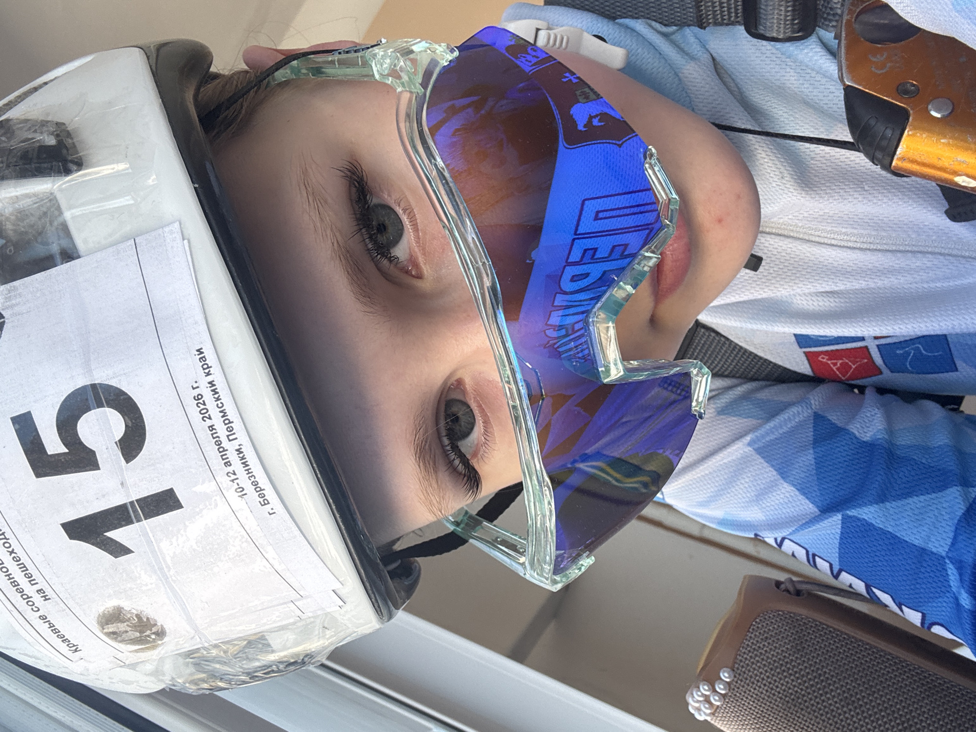 A close-up, rotated image shows a person wearing a white helmet with the number "15" on it. The text on the helmet indicates the location as "g. Bereznyki, Permskiy krai" and the date as "10-12 aprelya 2026 g." (April 10-12, 2026). The person is also wearing blue goggles. An orange device is visible in the upper right. The image orientation is rotated, making the objects appear sideways.