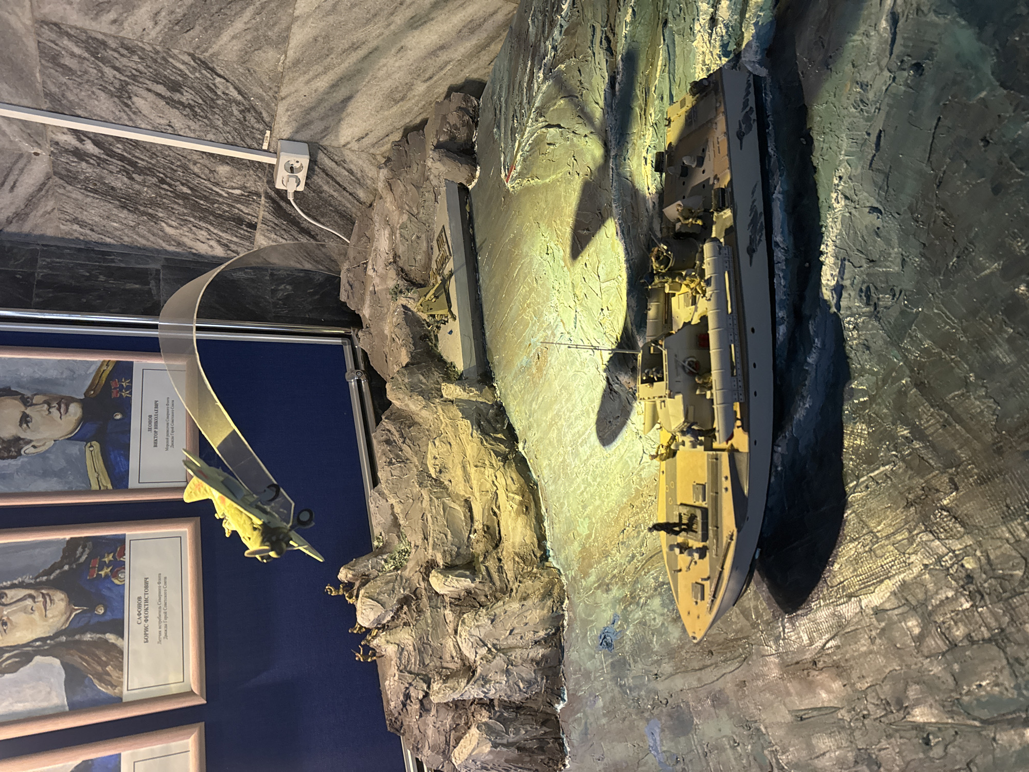 The image depicts a museum exhibit, likely located in Saint Petersburg, Russia, judging by the Cyrillic text on the portraits displayed. The exhibit showcases a diorama featuring a model warship navigating through textured, artificial cliffs or rocky terrain, presumably simulating a coastal environment. Above the warship is a model airplane, suspended by a clear plastic arc. To the left are framed portraits of individuals identified in Russian as, among other titles, "Twice Hero of the Soviet Union," accompanied by their names. The wall behind the portraits is a deep blue. An electrical outlet and track lighting are visible on the ceiling. The overall lighting in the diorama is warm, highlighting the details of the models and terrain.