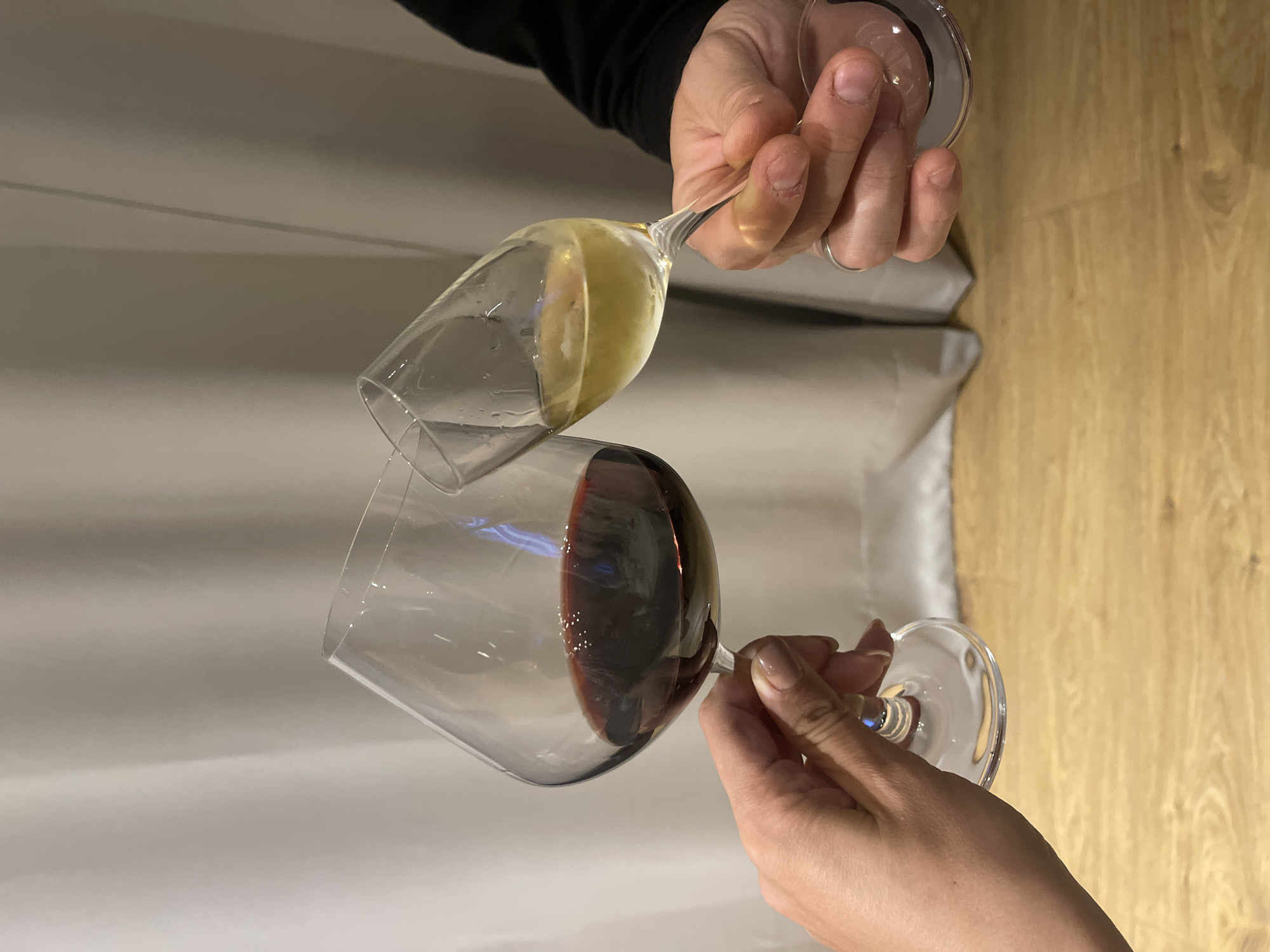 Two people are toasting with wine glasses filled with red and white wine. The background consists of a white surface and a wooden surface. The location in the city of Unknown, Unknown cannot be determined from the image.