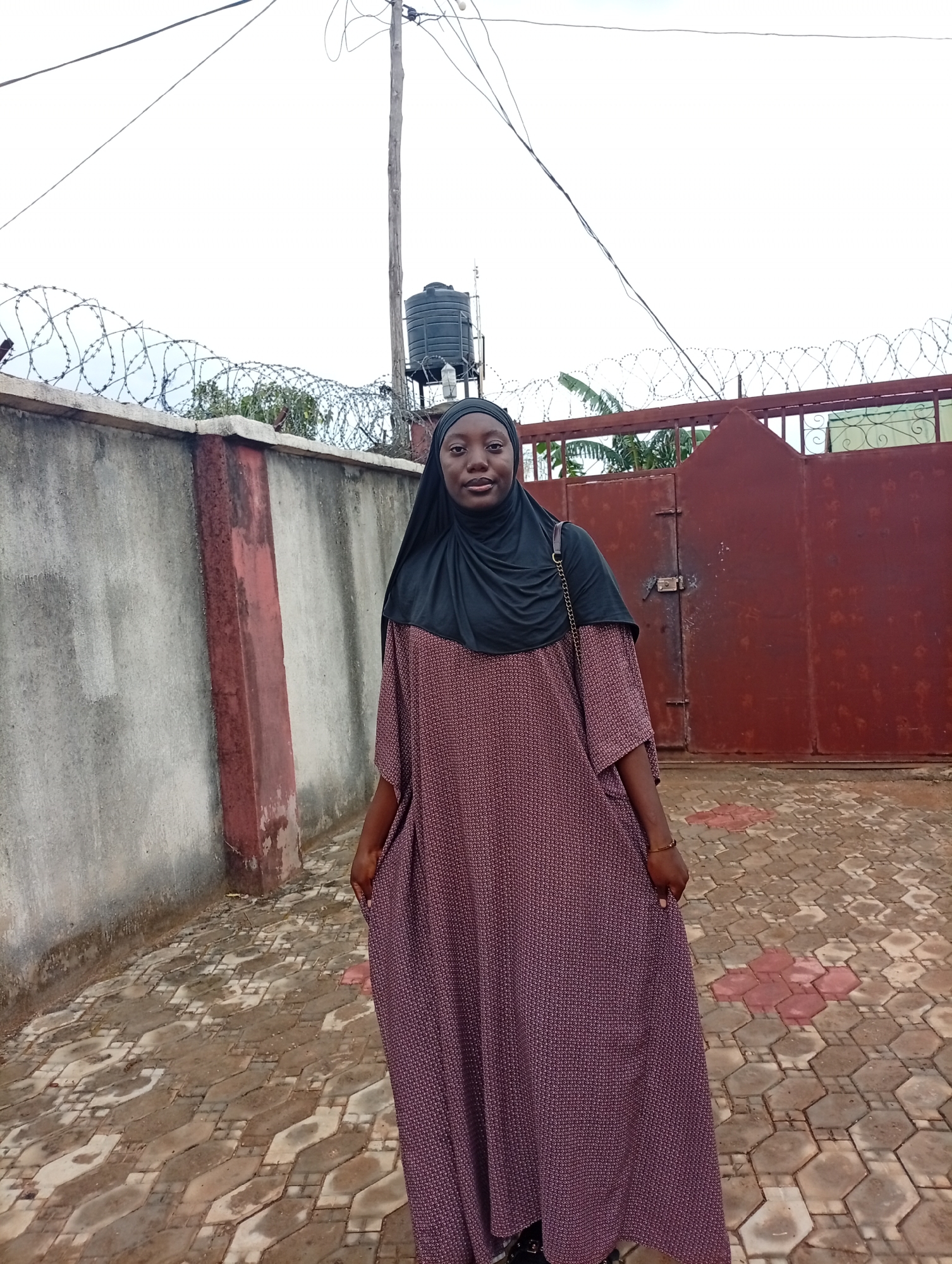 A woman wearing a black headscarf and a long, patterned dress stands in front of a reddish-brown metal gate and a concrete wall topped with barbed wire. The gate is locked. Behind the wall, a water tank is visible. The woman is facing the camera with her hands in her pockets. The ground is paved with interlocking bricks of varying colors. Overhead, power lines stretch across the scene. The location is Rido, Nigeria.