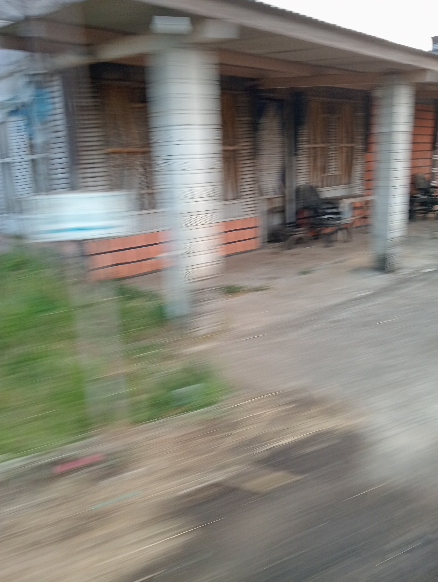 The image depicts a blurry outdoor scene of a building with a porch. The building appears to be a house or a small commercial establishment, with horizontal siding and several columns supporting a roof overhang. There are windows or openings with what appears to be blinds or shutters. On the porch, there are chairs, suggesting outdoor seating. The ground in front of the building is a paved or concrete surface, with some patches of grass to the left. The overall impression is of a somewhat weathered or perhaps abandoned property. The motion blur indicates the photo was taken from a moving vehicle. There are no people visible in the image.