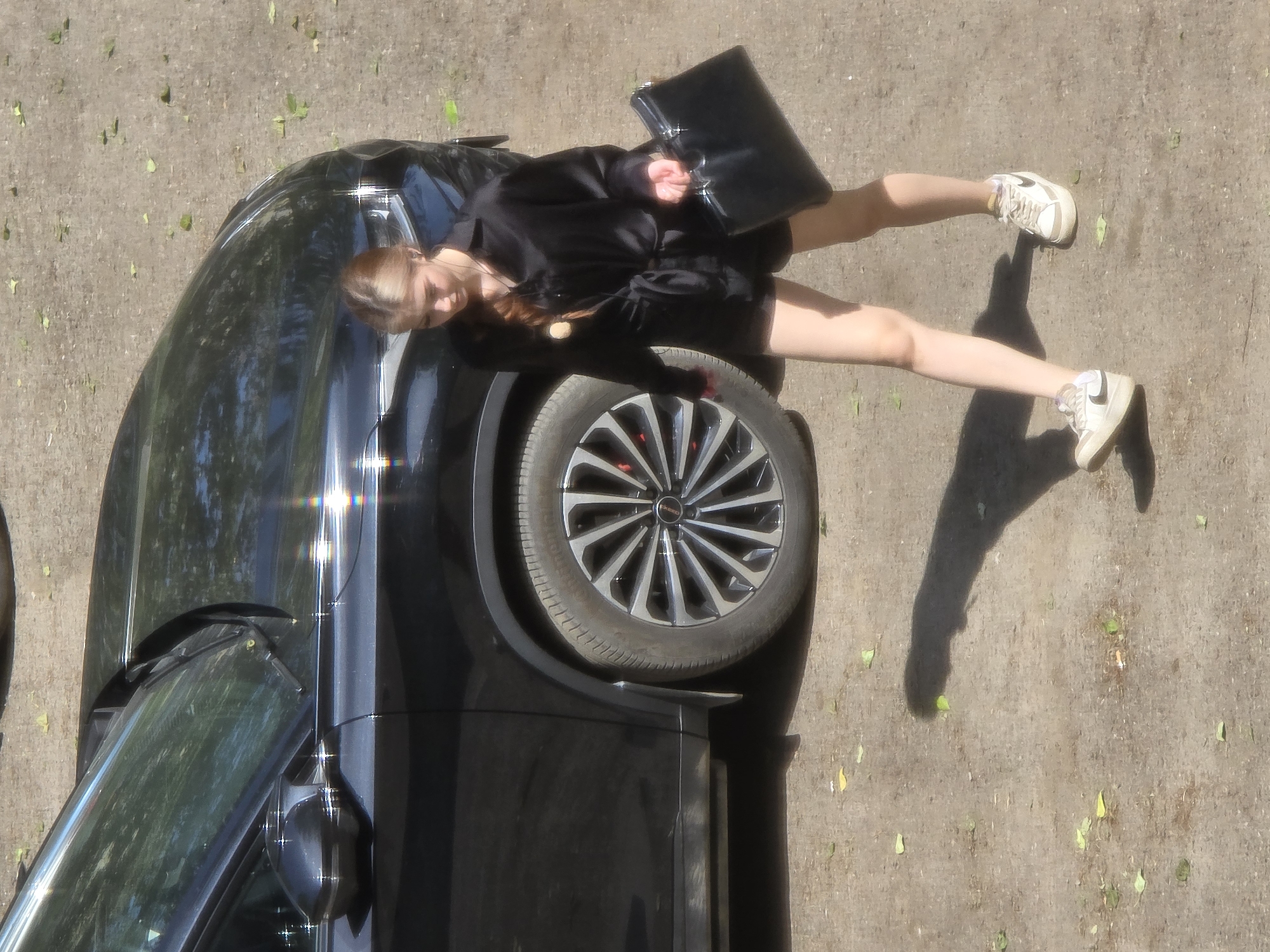 An overhead, medium shot captures a young woman with blonde hair in a braid walking next to a black car. She is wearing a black, short-sleeved collared shirt with a tie belt and black shorts. She carries a black, rectangular bag in her right hand and wears white sneakers. The car is parked on a dirt and leaf-strewn surface. Sunlight glints off the car's surface and casts a shadow of the woman onto the ground.