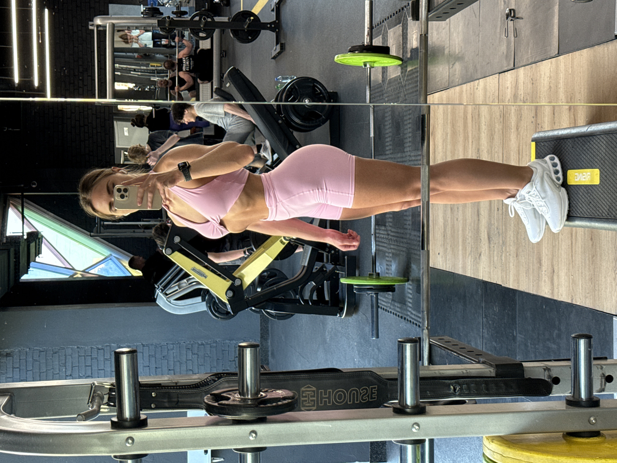 The image depicts a woman in athletic wear taking a selfie in a gym. She is wearing a pink sports bra and pink shorts, and white athletic shoes. She is standing on a black step with yellow markings. Her reflection is visible in a large mirror that dominates the left side of the frame. The gym appears to be well-equipped, with weight racks, barbells, weight plates (some green and yellow), and other exercise machines visible in the background. Other gym-goers are also visible in the reflection, some of whom are engaged in weightlifting. The overall environment suggests a typical fitness facility. There is no information in the image that indicates the location is in Saint Petersburg, Russia.