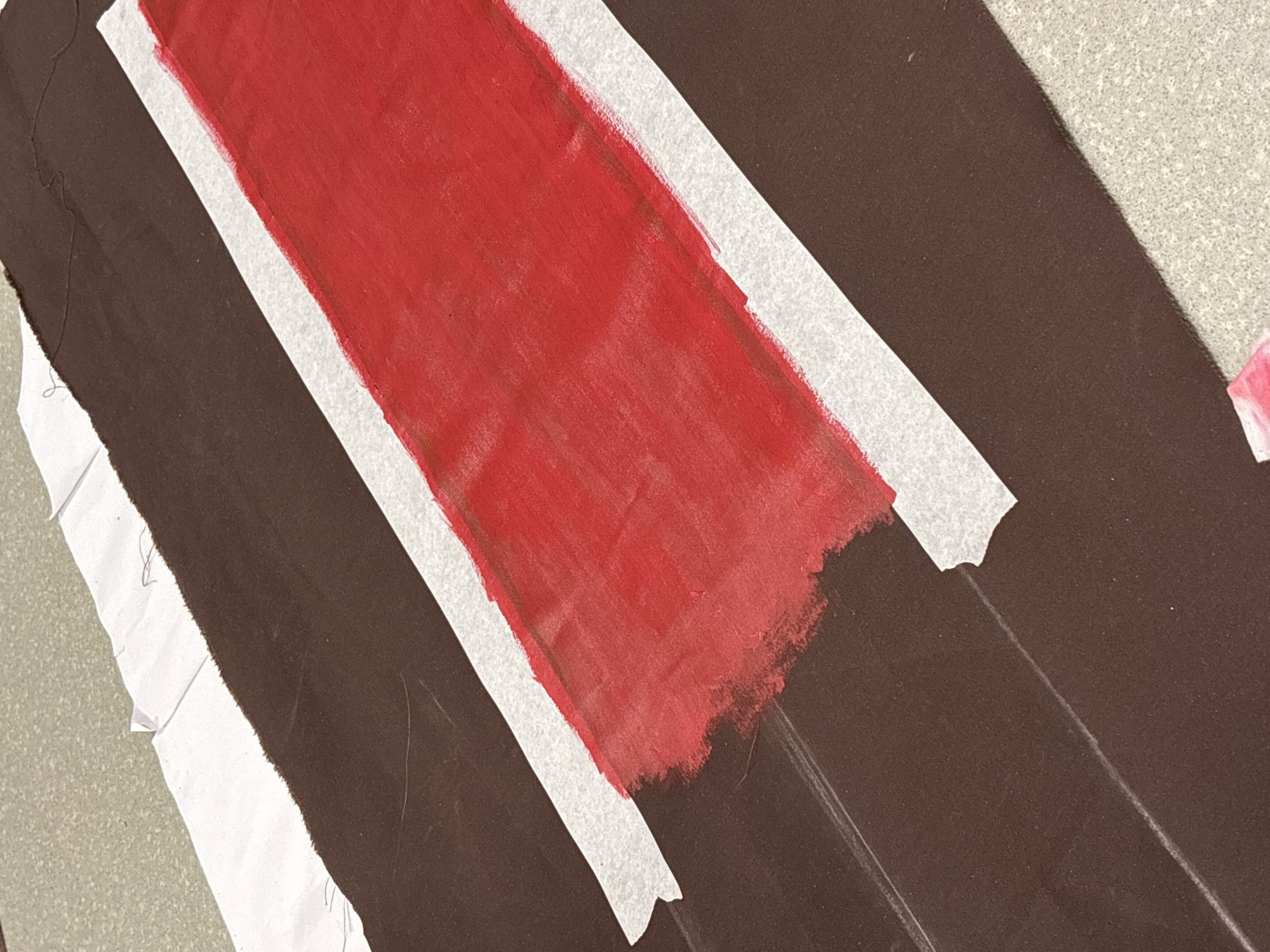 The image shows a piece of brown corrugated material with a red stripe painted on it. White masking tape borders the red stripe on both sides. The corrugated material and paint are on a surface.