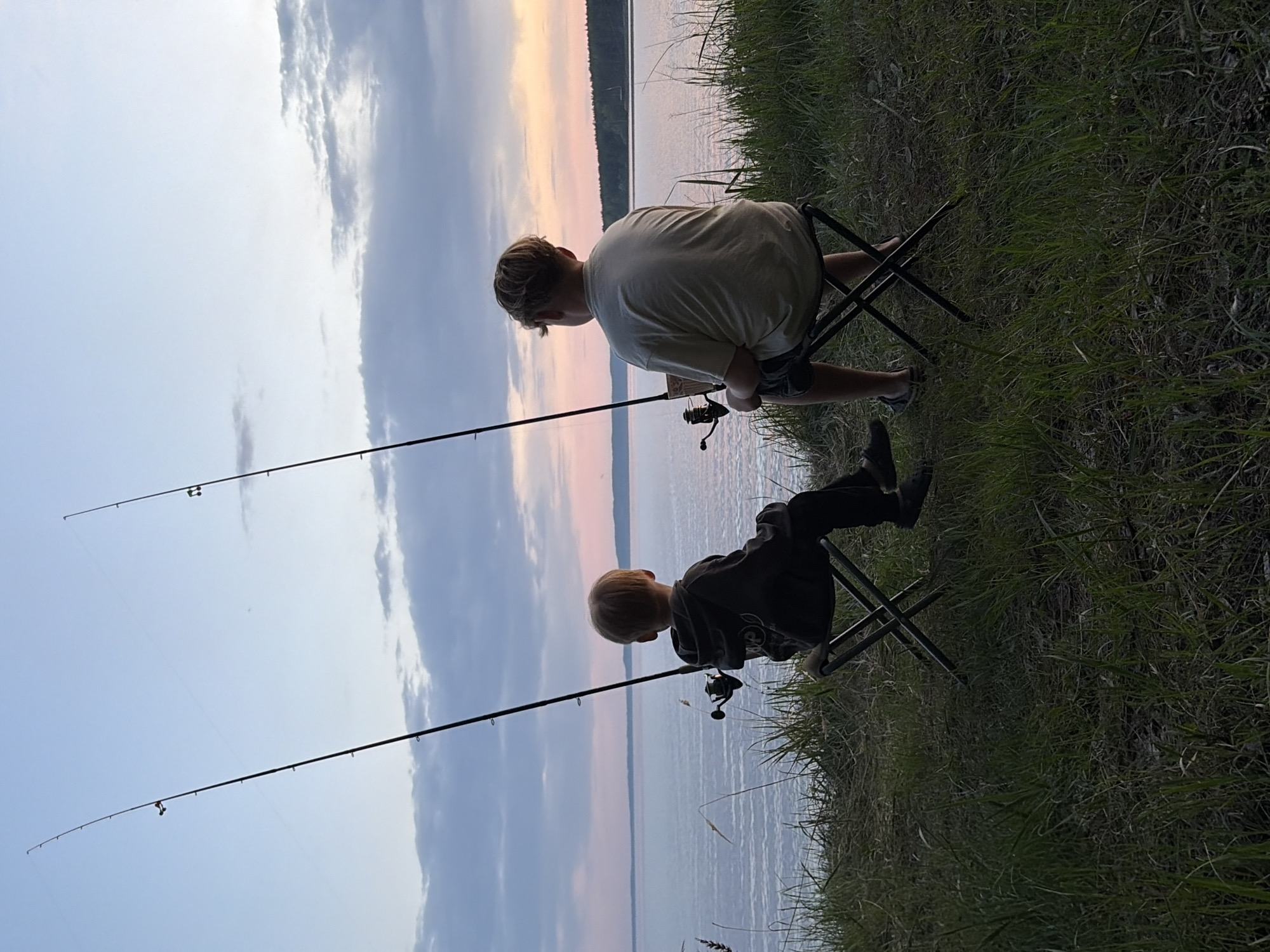 Two young people are fishing by a body of water. Both are sitting on folding chairs. Each holds a fishing rod, with the lines extending into the water. The body of water stretches to the horizon, where a dark treeline meets a pastel sky. The location is near Zabegaevo, Russia, based on other images from the same set. The foreground is grassy. The image is taken at eye level. The scene is likely at dusk, given the lighting.
