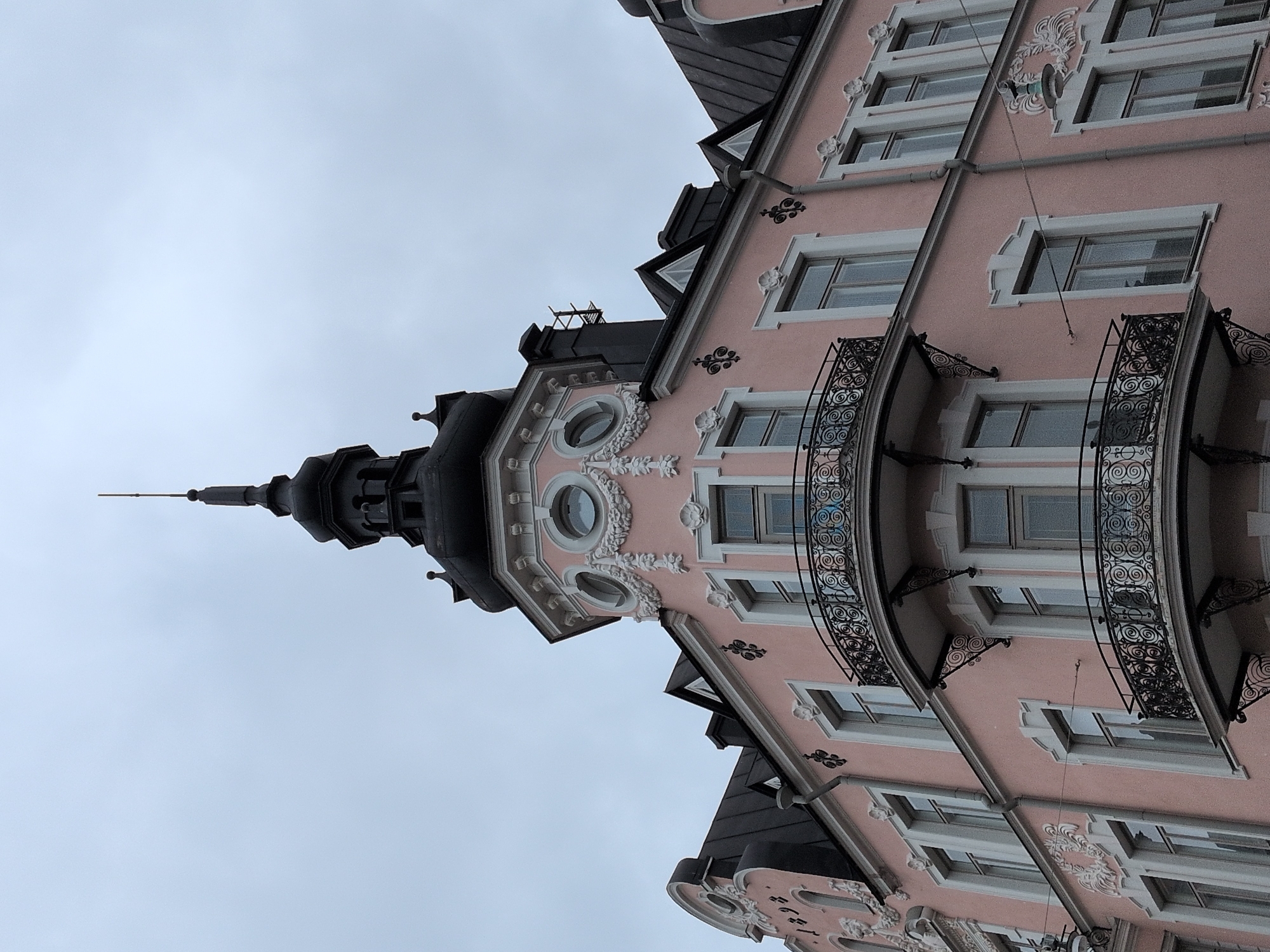 The image shows the Art Nouveau style building facade of the Huvilakatu 26 apartment building in Helsinki, Finland. The building is painted a pale pink color and features a prominent black domed turret with a spire. Decorative white plasterwork adorns the facade around the windows and balconies. The windows are tall and rectangular, with some featuring patterned glass. There are two wrought-iron balconies on the corner of the building, characterized by intricate scrollwork. The sky above is overcast and gray. The image appears to have been taken from street level, looking upwards at the building's facade.