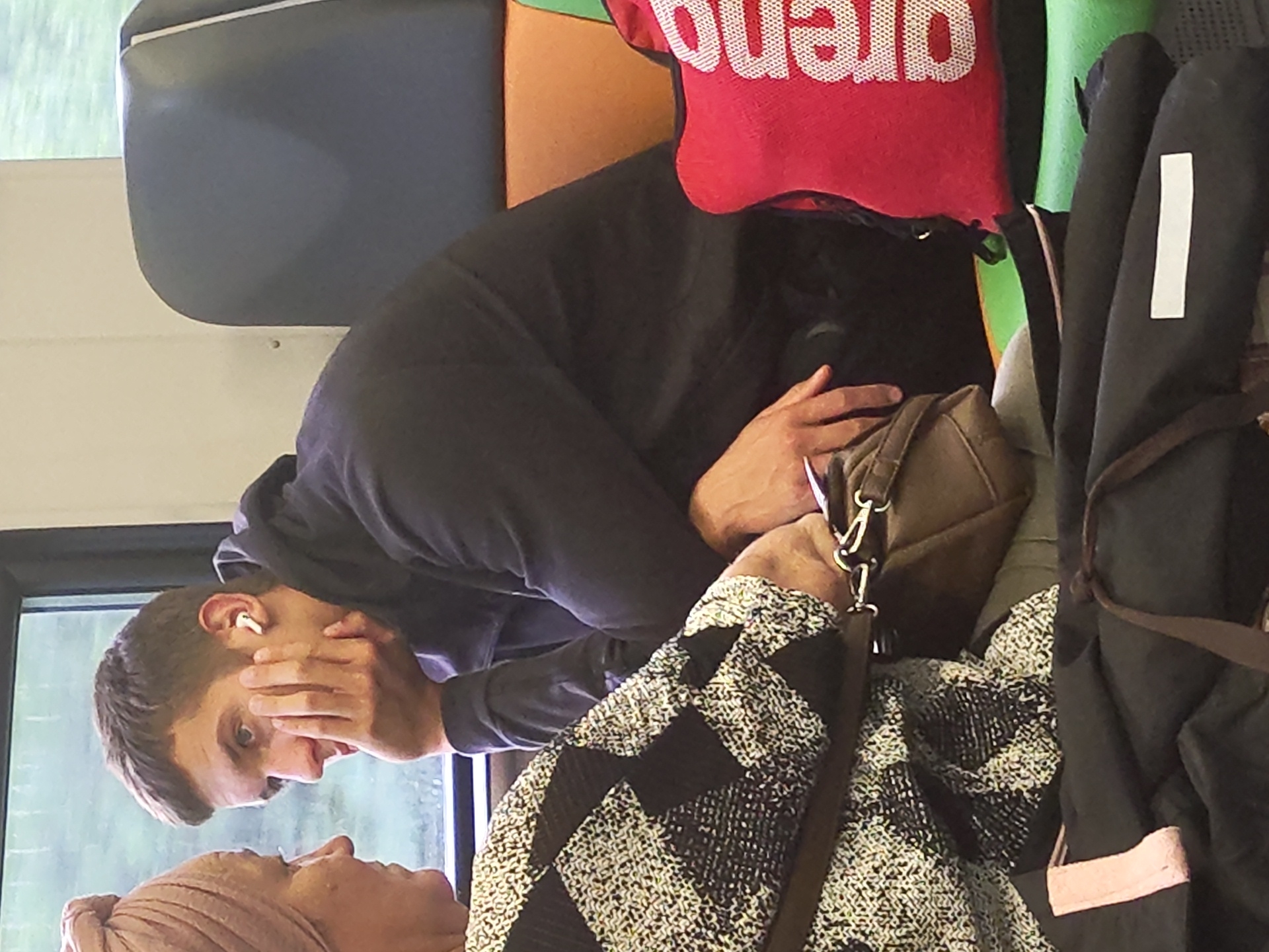 Two people are seated next to each other on a train. The person on the left, a woman, is wearing a headscarf and a black and white patterned top. Her left arm is resting on her lap. The person on the right, a man, is wearing a dark hooded sweatshirt and is looking towards the woman. He has a white earbud in his right ear and is resting his chin on his right hand. In front of the man is a red bag with white lettering that reads "arena". Various bags and objects are piled around the two individuals, suggesting a travel or commuting scenario. The background shows a train window with an out-of-focus view of greenery and a cloudy sky, consistent with a rural or suburban environment. The image was taken in the city of Rozhdestvenka, Russia.