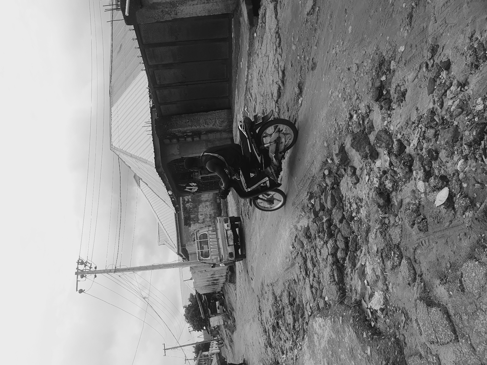 In Lokoja, Nigeria, a black-and-white photograph depicts a street scene. A man stands beside a parked motorcycle on a dirt road. The road surface is uneven and rocky. A small truck is parked further down the road. Overhead power lines run along the street. Buildings with corrugated metal roofs line the side of the road. The sky is overcast.