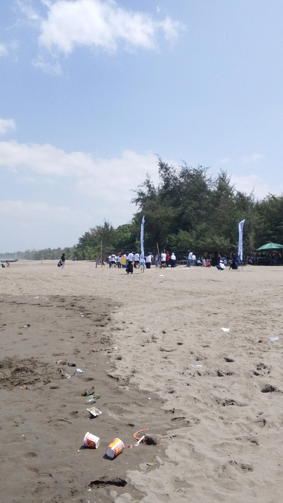 The image depicts a beach scene in Desa Pagandaran, Indonesia, under a partially cloudy sky. The foreground is dominated by wet sand with visible footprints and scattered litter, including plastic cups and wrappers. In the mid-ground, a group of people, mostly wearing white shirts, are gathered on the sandy beach. Several flags, two of which are white with blue lettering and logos, are planted in the sand near the group. A few trees and dense foliage line the background, with a small green canopy structure visible to the right. Further in the distance, a boat is visible on the left side of the frame. The overall impression is a daytime outdoor gathering on a coastal environment with evidence of recent human activity and some environmental pollution.