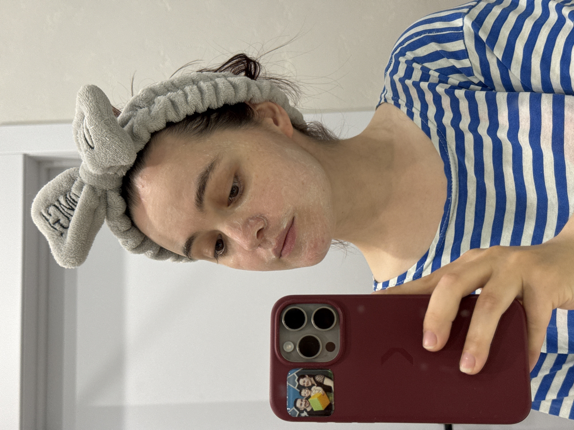 A young woman with a grey, fuzzy headband shaped like bunny ears is taking a selfie in a well-lit room, possibly a bathroom. She is wearing a blue and white striped t-shirt. Her face is partially covered in a white substance, possibly a face mask or cosmetic product. She is holding a dark red smartphone in her left hand, which has a small photo of a family attached to the case. The background is plain and light-colored, with a door visible on the left. There are no discernible landmarks to place this image specifically in Izhevsk, Russia, or any other city.