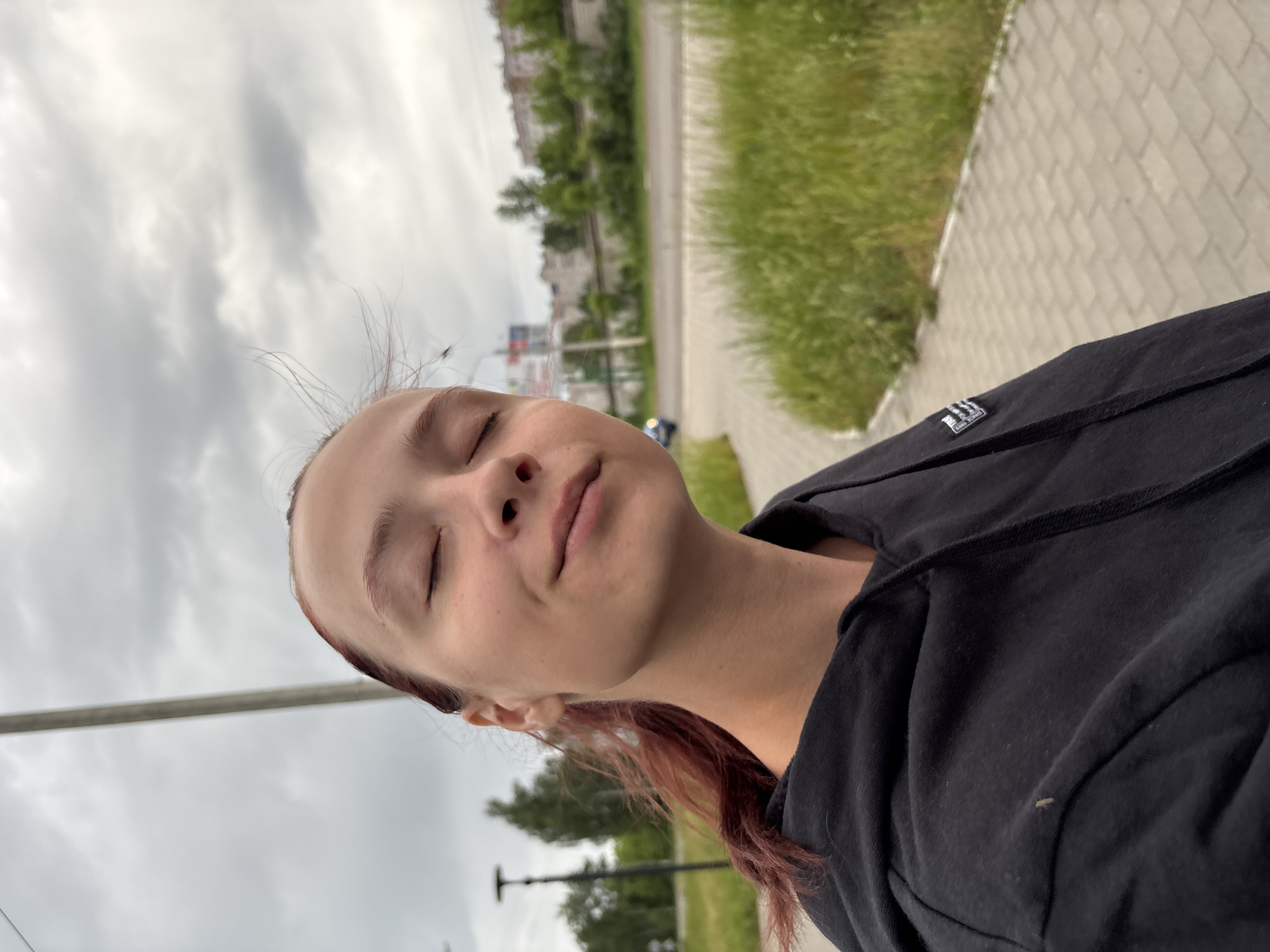 A woman stands outdoors on a paved pathway, likely in a park or urban area. She wears a black hooded sweatshirt and has her eyes closed, possibly enjoying the moment. Her hair is reddish-brown and pulled back. The background includes a road with green grass on one side and buildings in the distance. The sky is overcast. The specific location appears to be in Chaikovsky, Russia, based on visual indicators consistent with the city's environment.