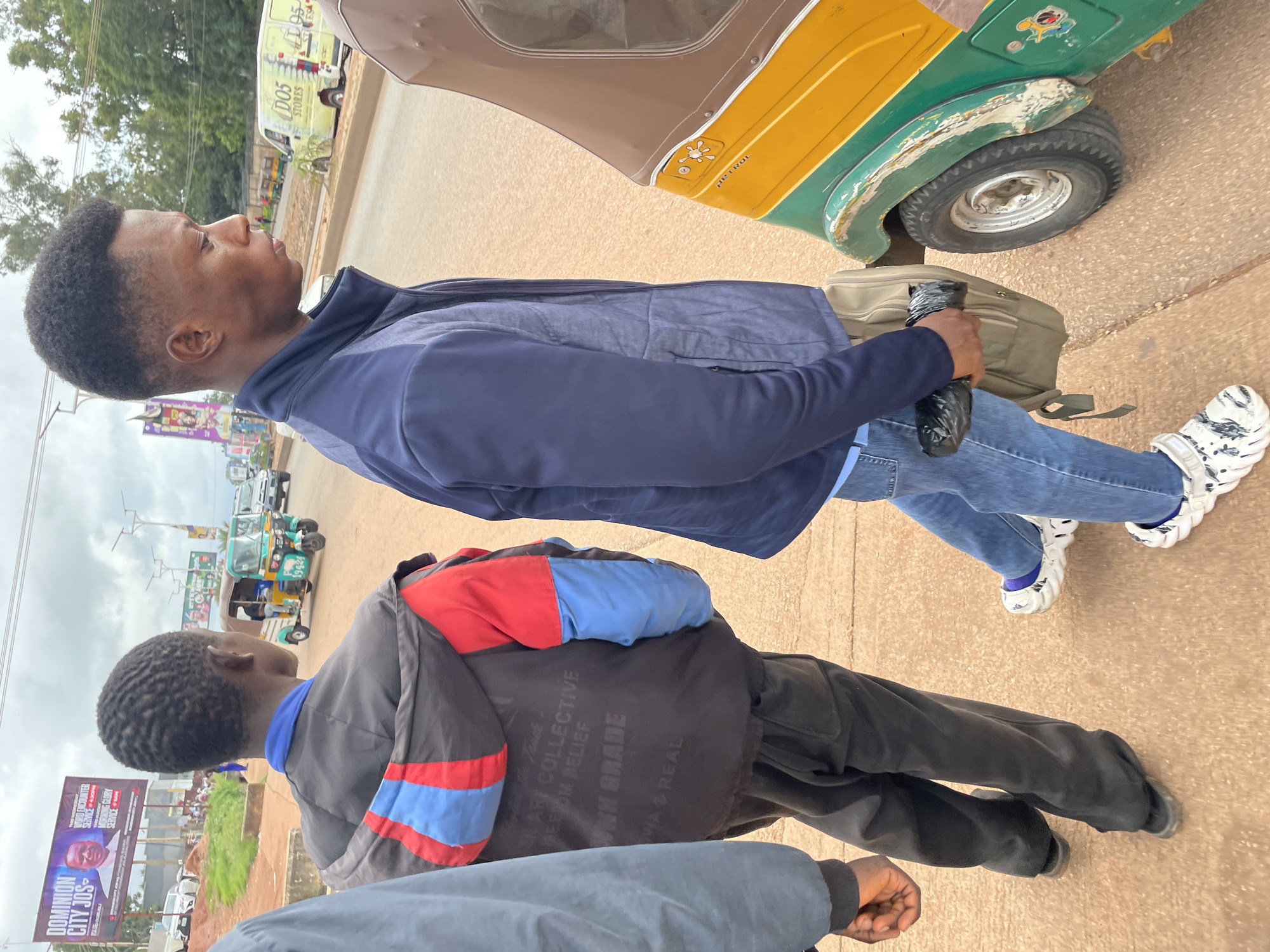 In Jos, Nigeria, three young men are walking along a paved road under an overcast sky. The man in the foreground, on the right, wears a blue puffer jacket over a blue shirt and blue jeans. He carries a tan backpack and a black plastic bag. His footwear consists of white, chunky-soled sandals with a black and white pattern. To his left, a young man in a black jacket with red and blue accents, black pants, and dark sandals is visible. A third man, partially seen on the far left, wears a grey long-sleeved shirt.
In the background, various elements indicate an urban setting in Jos. A green and yellow auto-rickshaw, a common mode of transportation in Nigeria, is parked on the right side of the road, its front tire visible. Further down the road, a light yellow van labeled "D05 STORES" is parked. Billboards are present on the left side of the street, including one with text in English and a portrait of a man, advertising "DOMINION CITY JOS." Other vehicles, including cars and more auto-rickshaws, are seen on the road and in the distance, suggesting moderate traffic. The ground is a reddish-brown dirt and asphalt mixture. Trees and buildings form the backdrop. The overall scene depicts a typical street view in Jos.