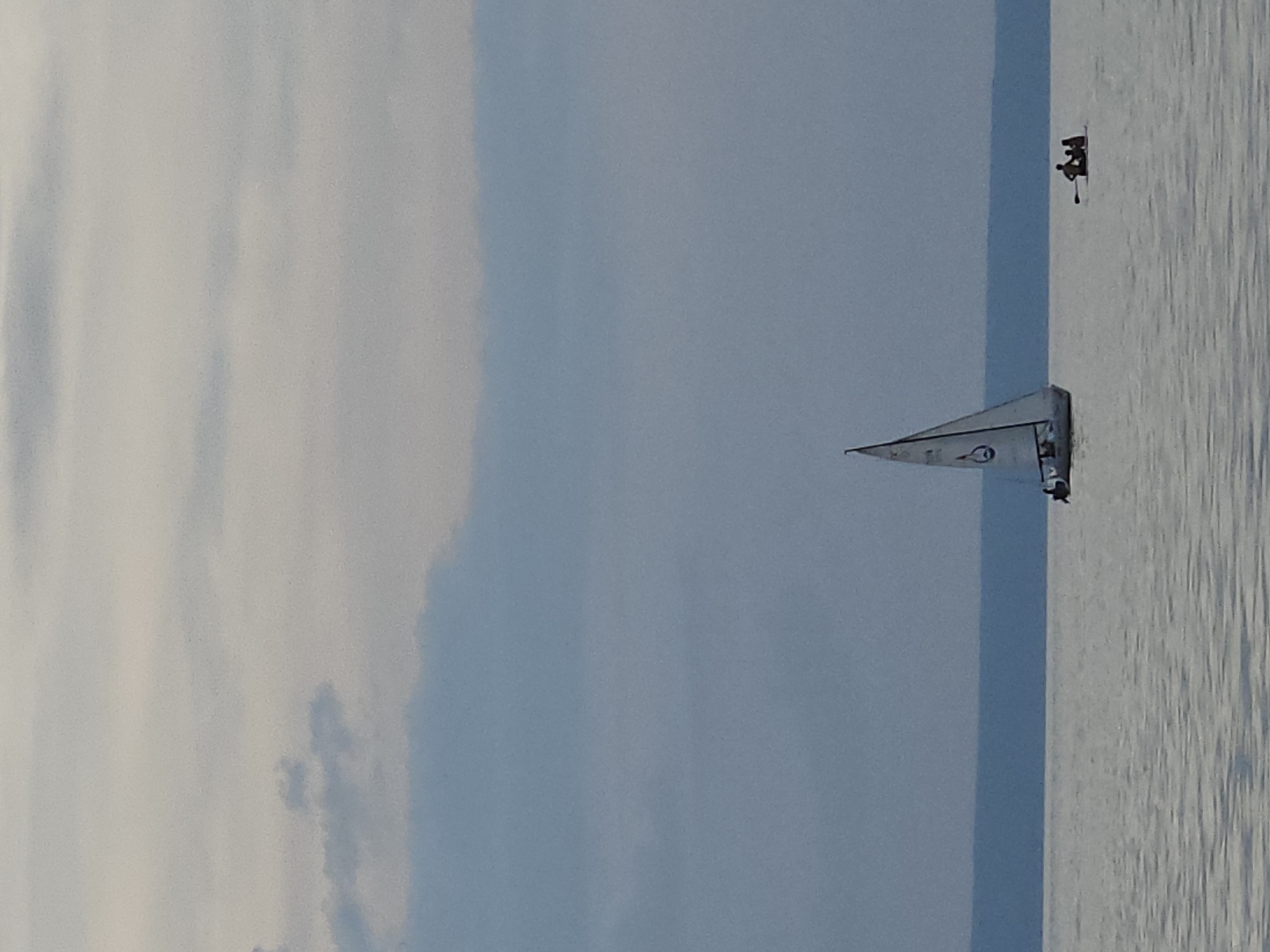 The image depicts a serene body of water, likely a lake or bay, under a cloudy sky. In the foreground, the water exhibits subtle ripples. A white sailboat with a black hull and a black outline of a circular symbol on its sail is positioned towards the left of the frame, appearing to be in motion. Several figures are visible on the sailboat. To the right of the sailboat, a small raft or paddleboard carries three silhouetted figures; one appears to be paddling. In the background, a distant, dark blue landmass stretches horizontally across the horizon. The sky is overcast with various shades of gray and light blue clouds. The overall impression is one of a peaceful, outdoor scene.