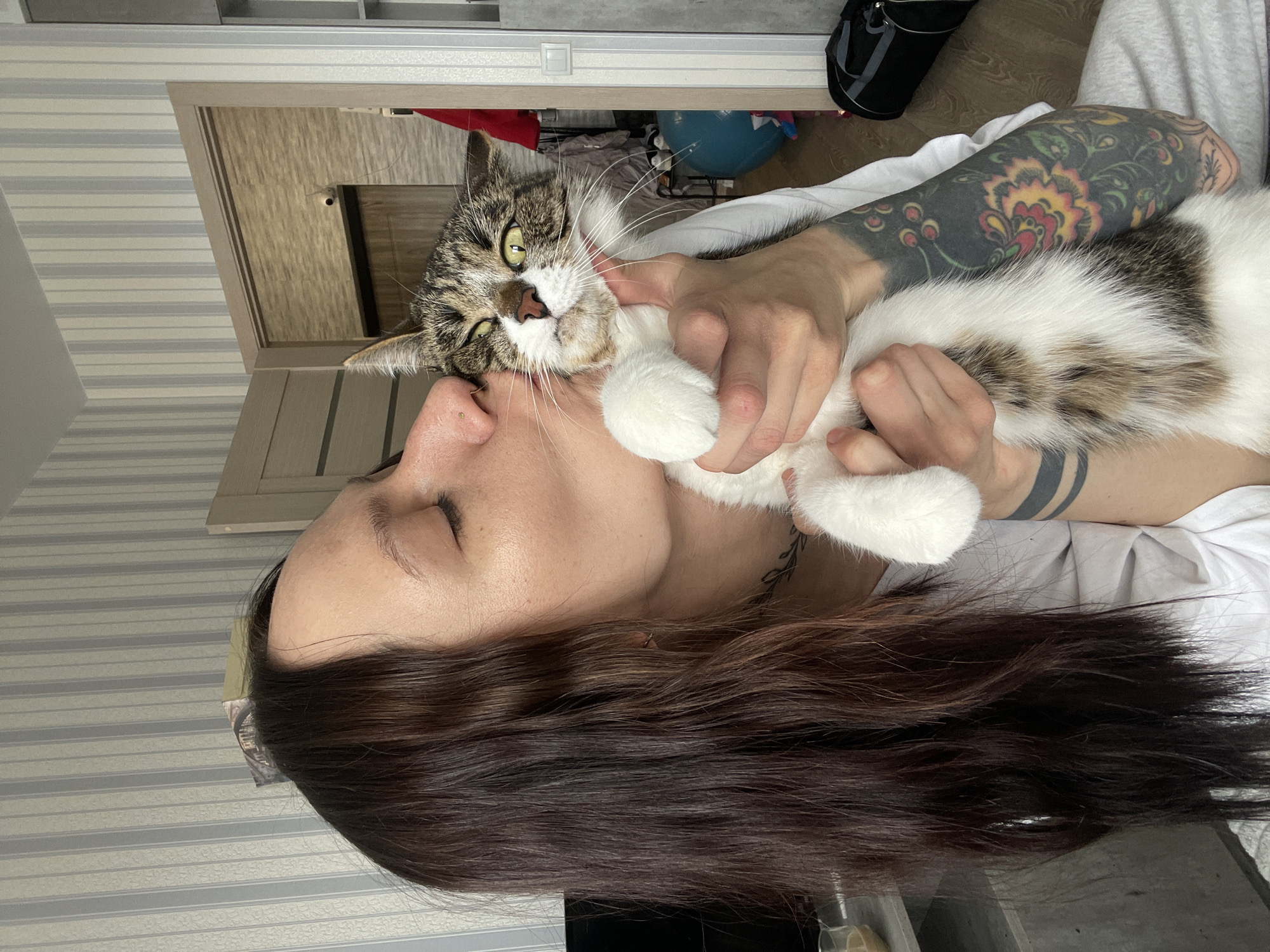A woman with dark hair is holding a tabby and white cat close to her face, possibly kissing it. The woman has her eyes closed. Her left arm is visible and has a large, colorful floral tattoo. She is wearing a white shirt. The cat is resisting or mildly annoyed, with its paws splayed open and a slight frown. They appear to be indoors, with a partially visible doorway and some household items in the background. There is no information available to indicate the location is Izhevsk, Russia.