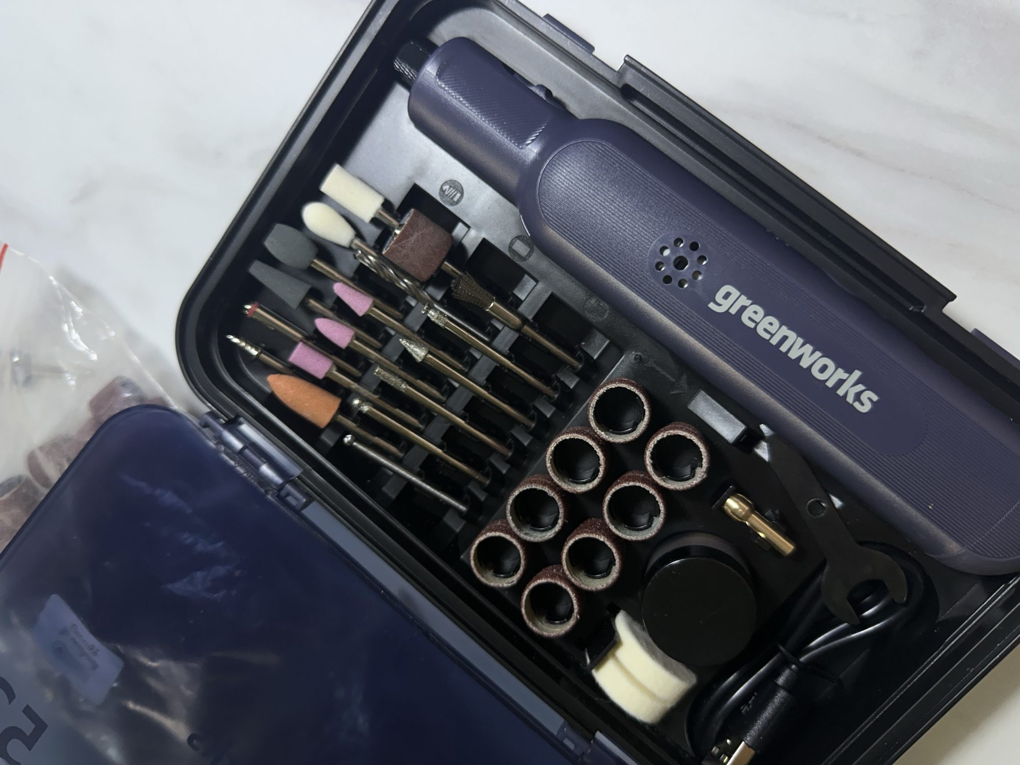 The image displays an open, dark-colored plastic storage case containing a compact rotary tool and numerous accessories. The main subject is a sleek, dark blue/purple handheld rotary tool, prominently featuring the "greenworks" logo in white text on its side. It has a textured grip and a perforated section near the logo, likely for ventilation.
The case's interior is meticulously organized with custom-fit compartments. To the left of the tool, there's an array of interchangeable bits, including various shapes and materials for grinding, sanding, and polishing. These include conical pink and orange bits, cylindrical brown bits, gray and white abrasive stones, and white felt-like polishing buffers. Several metallic engraving or cutting bits with fine points are also visible. Below these are cylindrical sanding bands, neatly stacked in rows.
Other accessories in the case include a small black wrench, a gold-colored collet, a coiled black USB charging cable, and a small black circular container. A clear plastic bag, partially visible on the far left, contains additional small items. The case's translucent dark blue lid is open and partially visible in the lower-left corner. The background appears to be a light-colored, possibly white or marble-patterned surface, suggesting an indoor setting. The items appear new and well-organized, indicating a complete kit ready for use. No specific location cues for Kuda, Russia, are discernible from the image.