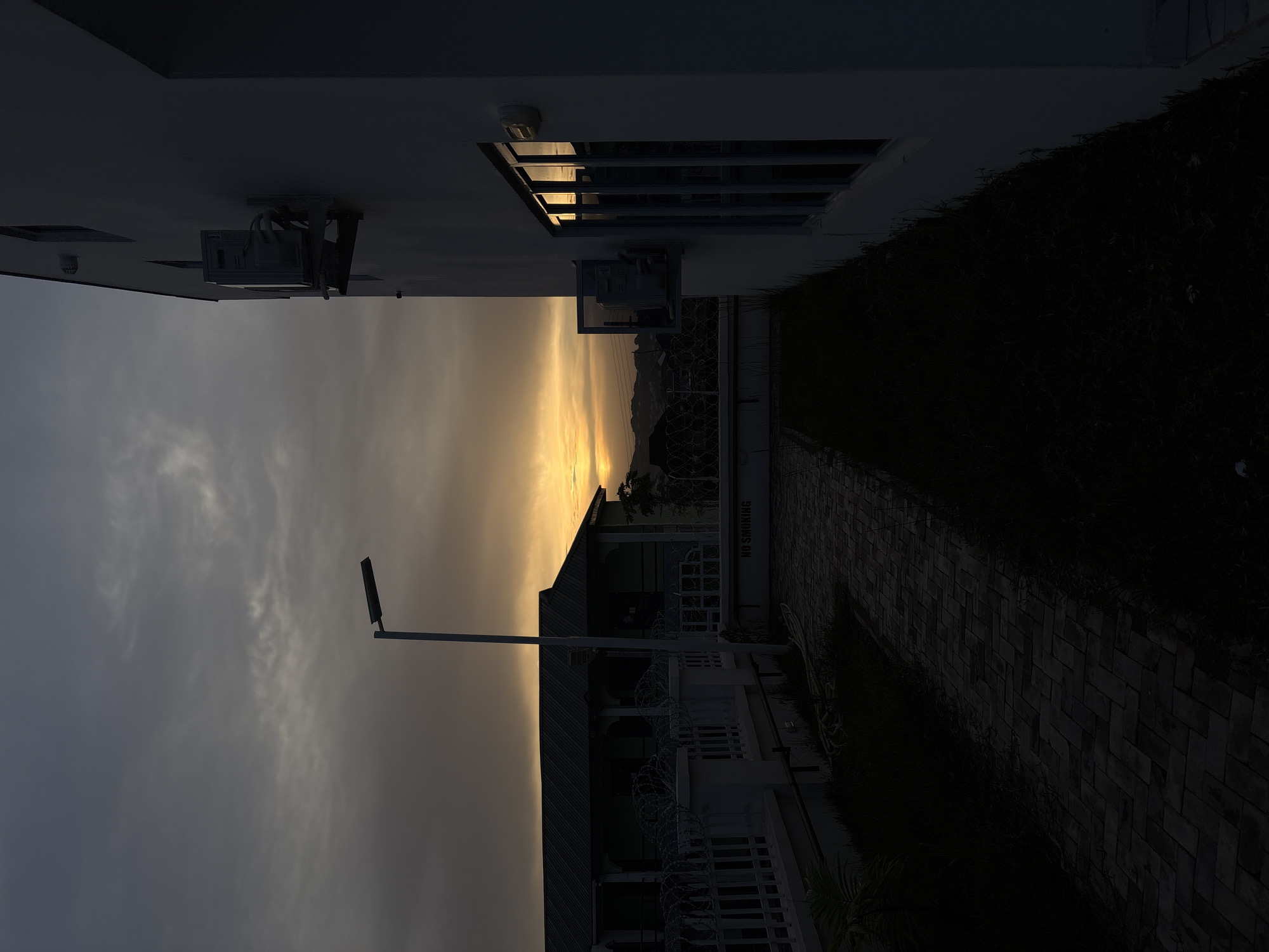 This image, set in Bukuru, Nigeria, depicts an outdoor scene during either sunrise or sunset, characterized by a dramatic golden hour glow. The sky, a dominant feature, transitions from a soft, cloudy grey-blue in the upper regions to a brilliant orange and yellow hue along the horizon, suggesting the sun's position just out of frame or behind a distant landmass.
In the left foreground, a modern building stands as a dark silhouette against the bright sky. Its facade, likely white, features an outdoor air conditioning unit and a rectangular window that reflects the intense light. A white railing topped with barbed wire runs along the building's base. Another building with a dark roofline is visible further back, also partially obscured by shadows. A tall street lamp with a flat, rectangular light fixture is prominently silhouetted in the mid-ground.
To the right of the buildings, a paved pathway made of dark bricks laid in a herringbone pattern stretches towards the background. Dark green foliage and grass line the pathway and the base of the structures. A distinct white sign or wall segment near the pathway clearly displays the text "NO SMOKING". In the far distance, faint outlines of hills or mountains are discernible beneath the illuminated horizon. The overall scene is serene and emphasizes the contrast between the vibrant sky and the shadowed foreground elements.