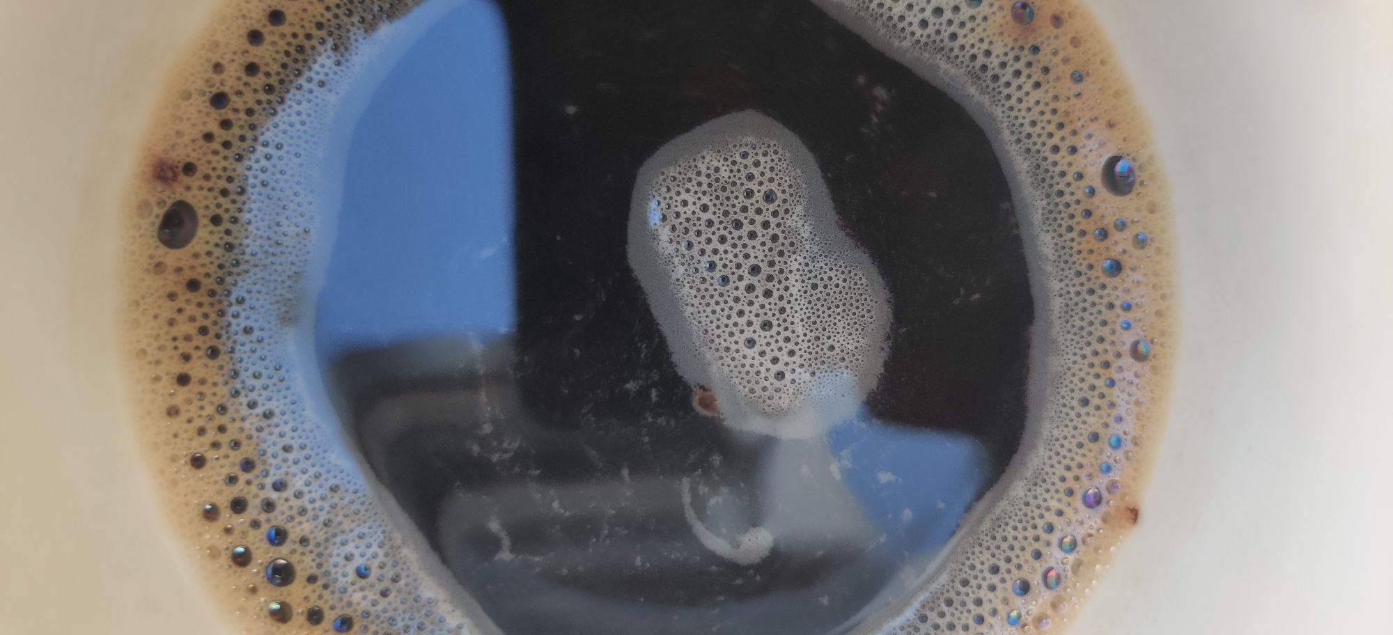 A close-up, overhead shot captures the foamy surface of a cup of dark coffee. The coffee has a rich brown color, and the foam is a lighter tan, creating a distinct border around the dark liquid. Small, iridescent bubbles dot the foam, reflecting hints of purple and blue. A larger, isolated patch of foam with denser bubbles sits in the center of the coffee. The coffee itself appears dark and reflective, showing a blurred blue and white reflection, possibly of a window or sky. The rim of the white ceramic cup is visible at the bottom and sides of the frame. The overall impression is one of a fresh, hot beverage. There are no people or identifiable landmarks in the image. The location in Rizunenkove, Ukraine, is not discernible from the visual information provided.