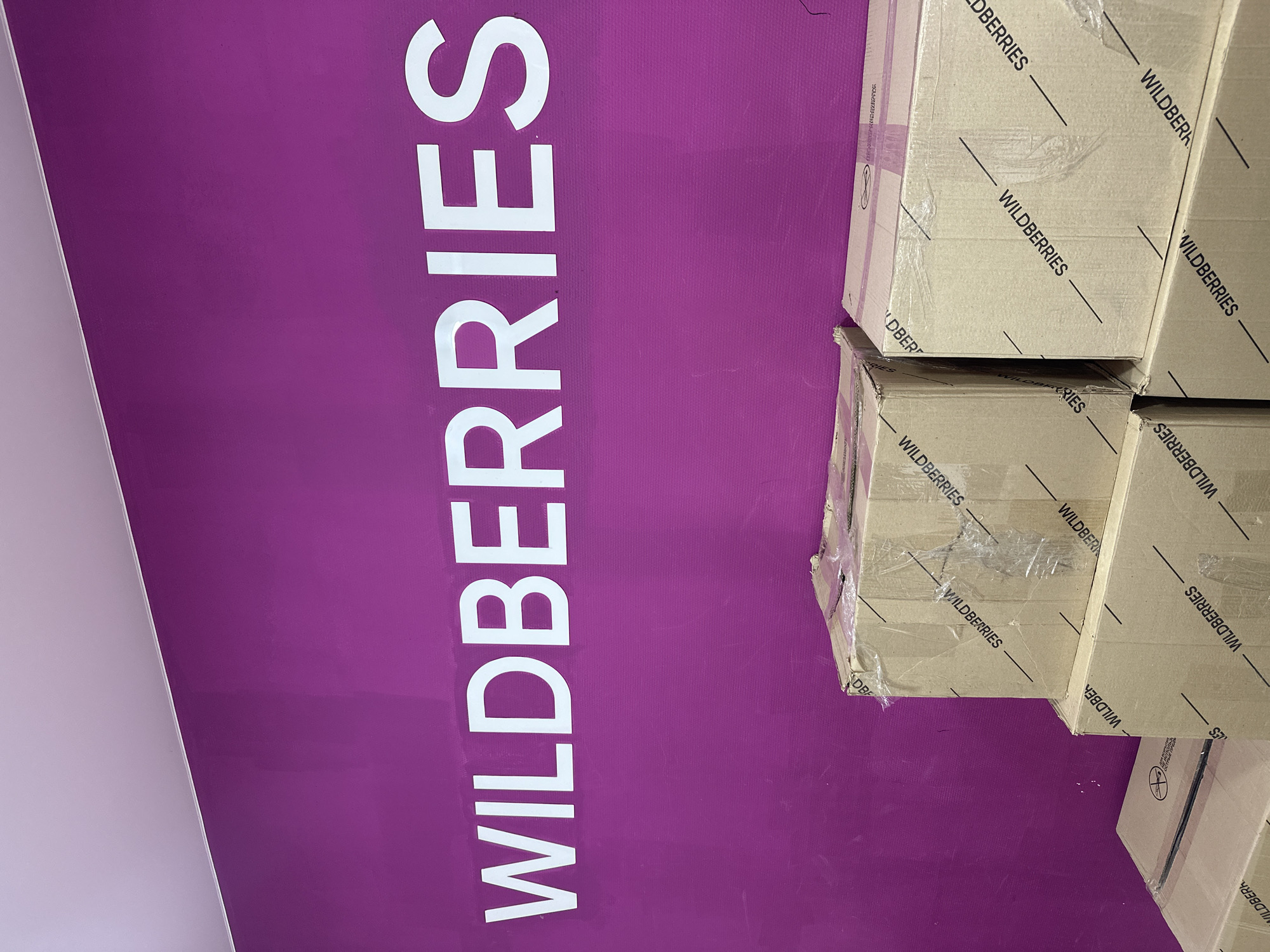 A close-up, eye-level shot shows a magenta wall with large white letters spelling out "WILDBERRIES" in the upper half of the frame. The wall has a slightly textured surface. Below the lettering, stacks of cardboard boxes are visible, most of which are taped shut and have "WILDBERRIES" printed repeatedly on them in black lettering. The boxes appear to be part of a shipment or storage. The lighting suggests an indoor environment, possibly a warehouse or retail space, with no people or animals present. The image does not provide specific indicators for its location within Novoaltaysk, Russia, beyond the presence of the "WILDBERRIES" branding, which is associated with an e-commerce company.