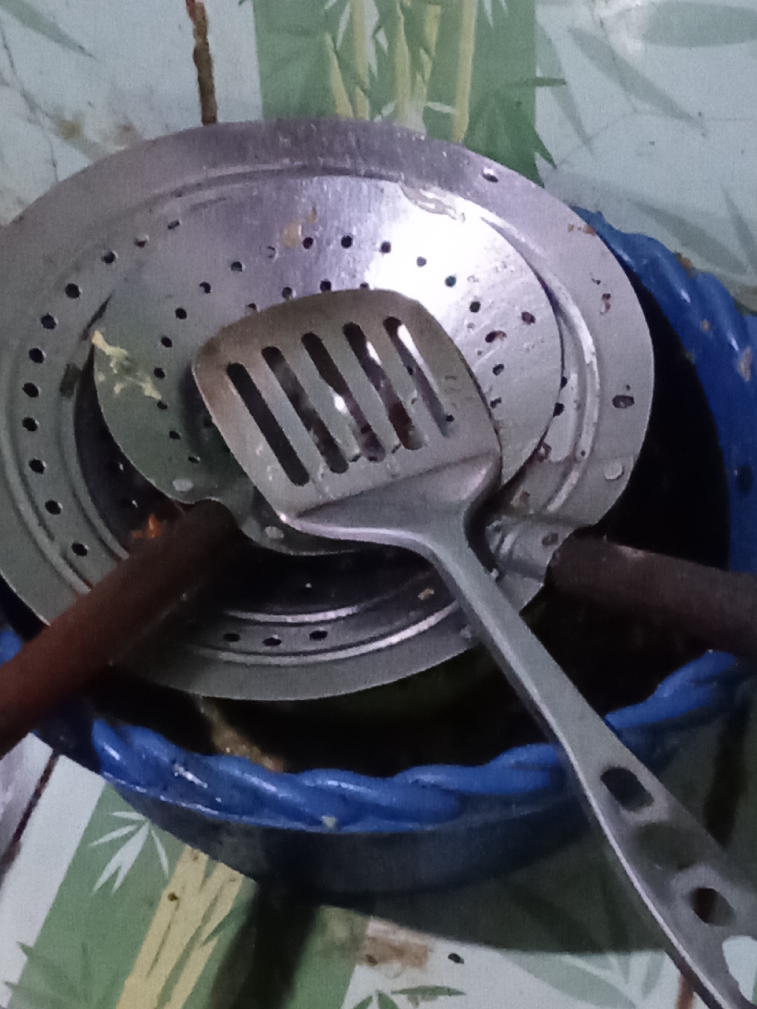 The image shows kitchen utensils, including a slotted spatula and two circular metal strainers or colanders, stacked inside a blue plastic container. The utensils appear to be used, with some residue visible on them. The background consists of a wall with a green and white bamboo-like pattern. The scene is dimly lit, suggesting an indoor setting. The image has been taken from a high angle, looking down at the objects. There are no people visible in the image. The overall impression is of a domestic kitchen environment. Given the limited information, it's impossible to determine the specific location within Pemalang, Indonesia, beyond it being an indoor kitchen.