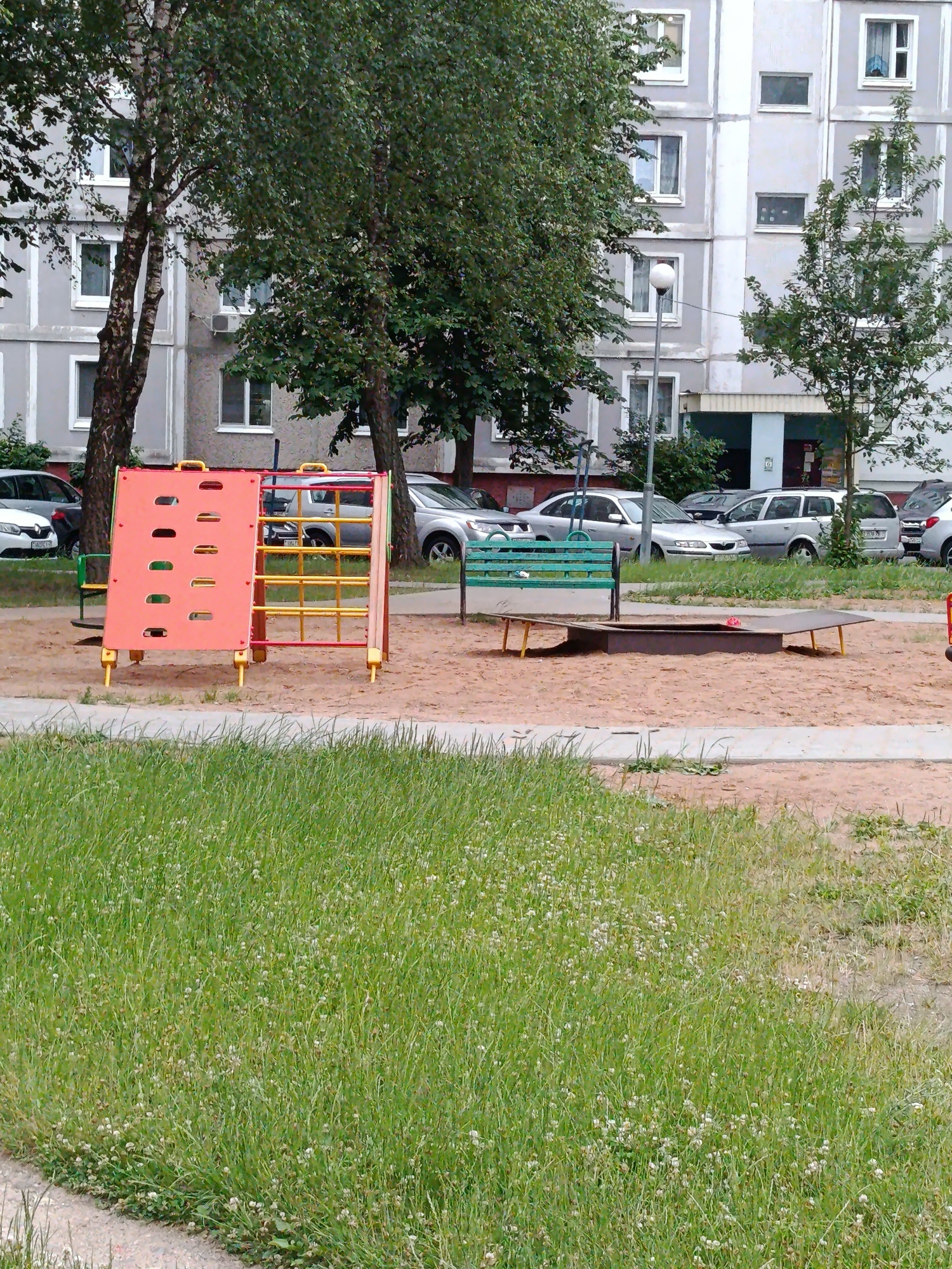 This image depicts a children's playground situated in front of a multi-story apartment building in Minsk, Belarus. The playground features a bright orange and yellow climbing structure with numerous handholds and a ladder. Adjacent to it is a green wooden bench placed near a sandpit. A small red object, possibly a toy, is visible in the sandpit.
The scene is set outdoors, with lush green grass covering the foreground and surrounding area. Several trees with dense foliage provide shade. In the background, the gray apartment building dominates the scene, with multiple windows and balconies visible. A street lamp stands near the building. Several cars, including sedans and an SUV, are parked in front of the building, suggesting it's a residential area.
The lighting suggests it is daytime, possibly overcast as there are no strong shadows. The overall atmosphere appears calm and ordinary, typical of a residential neighborhood. No people are visible in the image. The image lacks any discernible text, apart from what might be on the car license plates, which are too blurry to read.