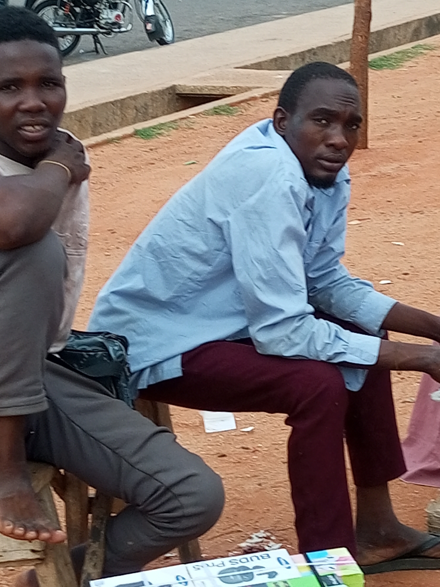 In Gombe, Nigeria, two men are seated on wooden stools outdoors. The man on the left, wearing a light-colored shirt and gray pants, is partially visible, with his right arm resting on his leg. He has a bracelet on his wrist. The man on the right wears a light blue long-sleeved shirt and maroon pants. He is looking towards the viewer, with a slight beard. A motorcycle is visible in the background. The ground is covered in red dirt, and a concrete structure with a drainage channel runs along the edge of a sidewalk. Various objects, including a dark bag and what appear to be packaged goods or books, are scattered around the men. The overall scene suggests a casual outdoor setting, possibly a marketplace or a roadside resting area.