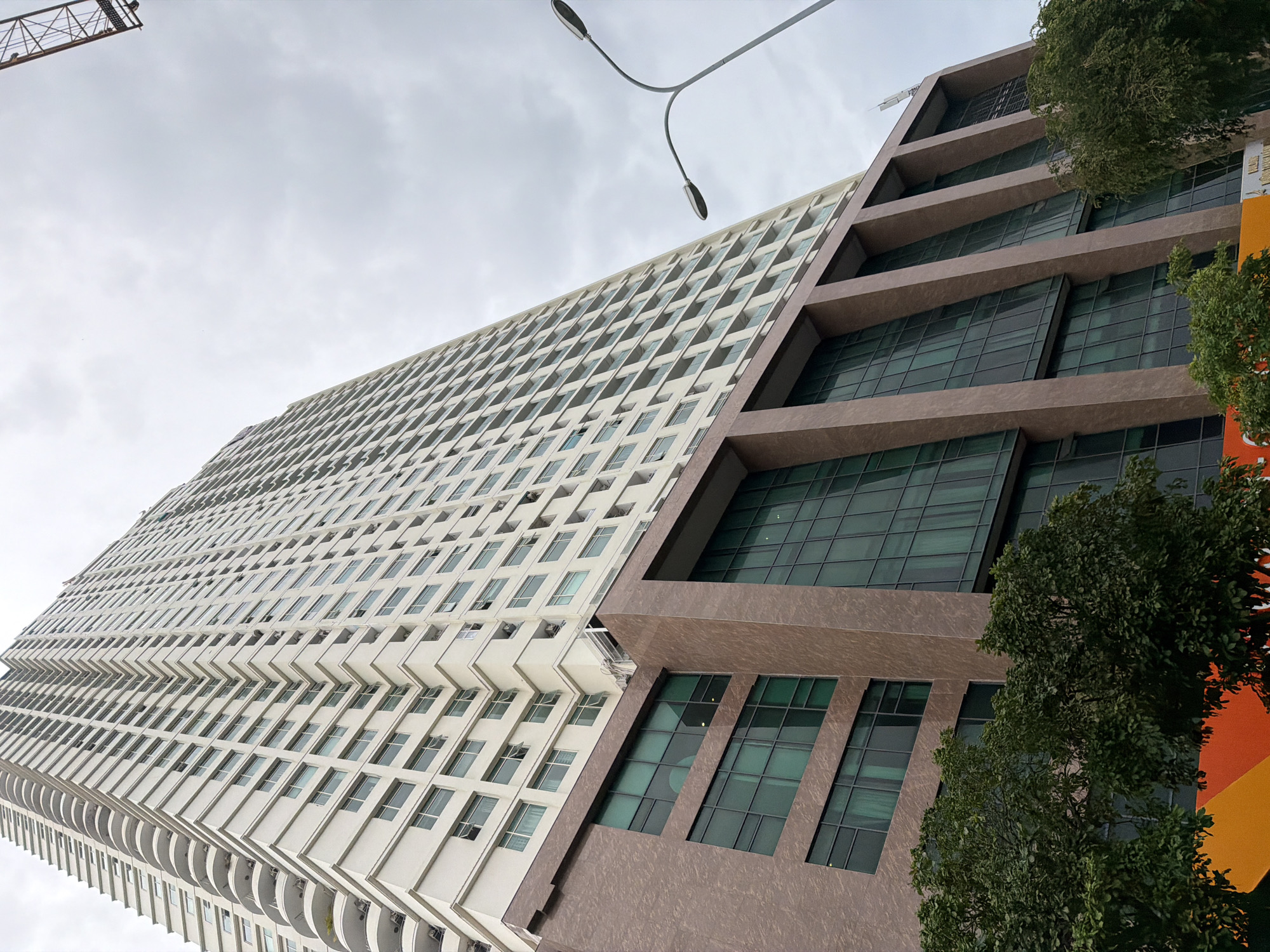 The image depicts a low-angle, wide shot of a tall, modern apartment building in Nha Trang, Vietnam. The building is primarily white with a repetitive pattern of windows and balconies. The lower section of the building has a brown, textured facade with large, dark green windows. Trees with dense green foliage are visible in the foreground, partially obscuring the base of the building. In the upper right corner, a portion of a construction crane is visible against a cloudy sky. A street lamp with two lights is positioned to the right of the building. The scene appears to be outdoors on an overcast day.
