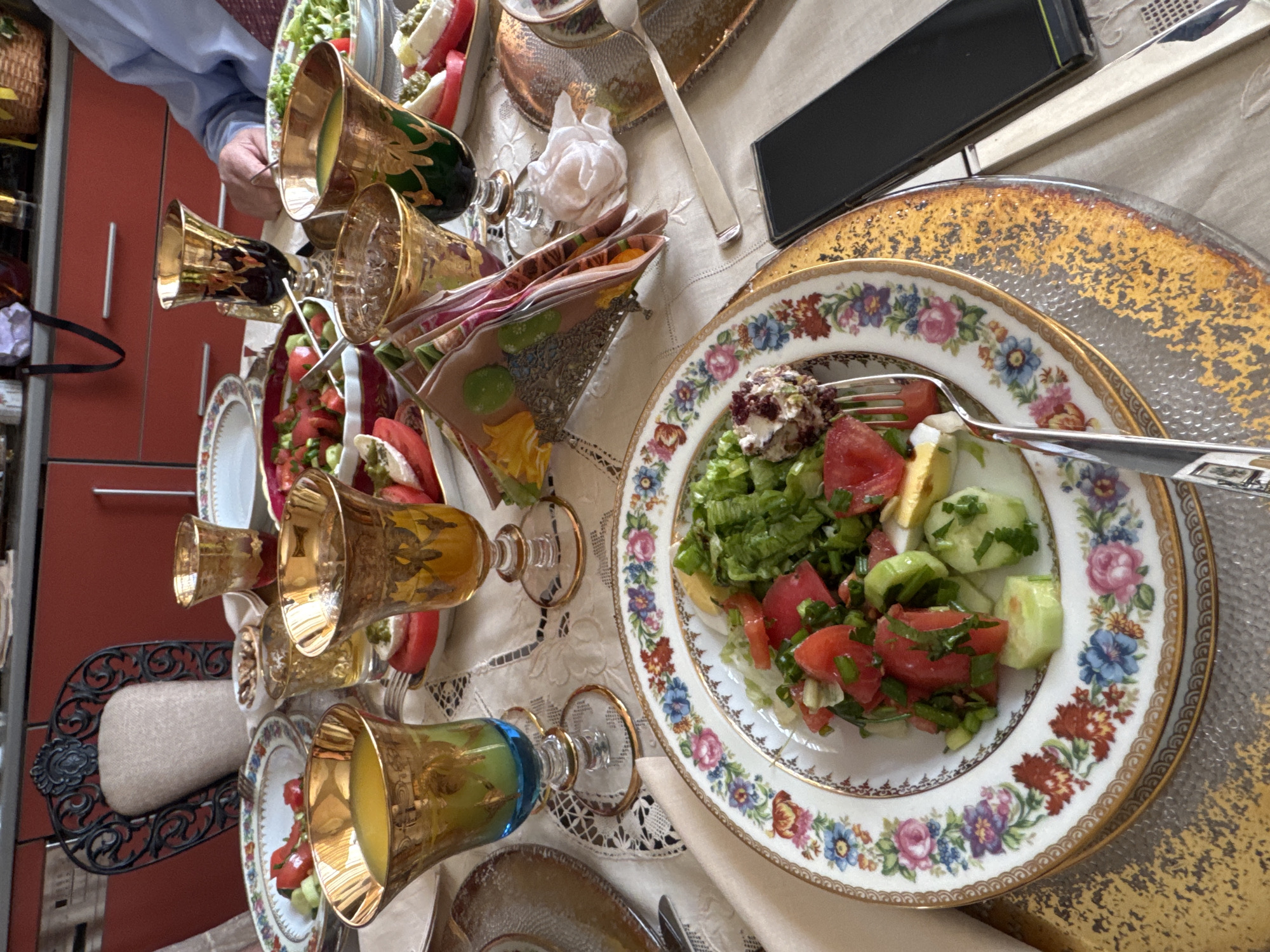 The image shows a table setting, likely indoors, featuring fine china plates with floral patterns, glassware with gold accents, and a salad. A person's arm and hand are visible on the left side of the frame. A fork is in use, piercing the salad. A mobile phone is placed on the table. The setting suggests a formal occasion. The location in Plovdiv, Bulgaria, cannot be determined from the image.