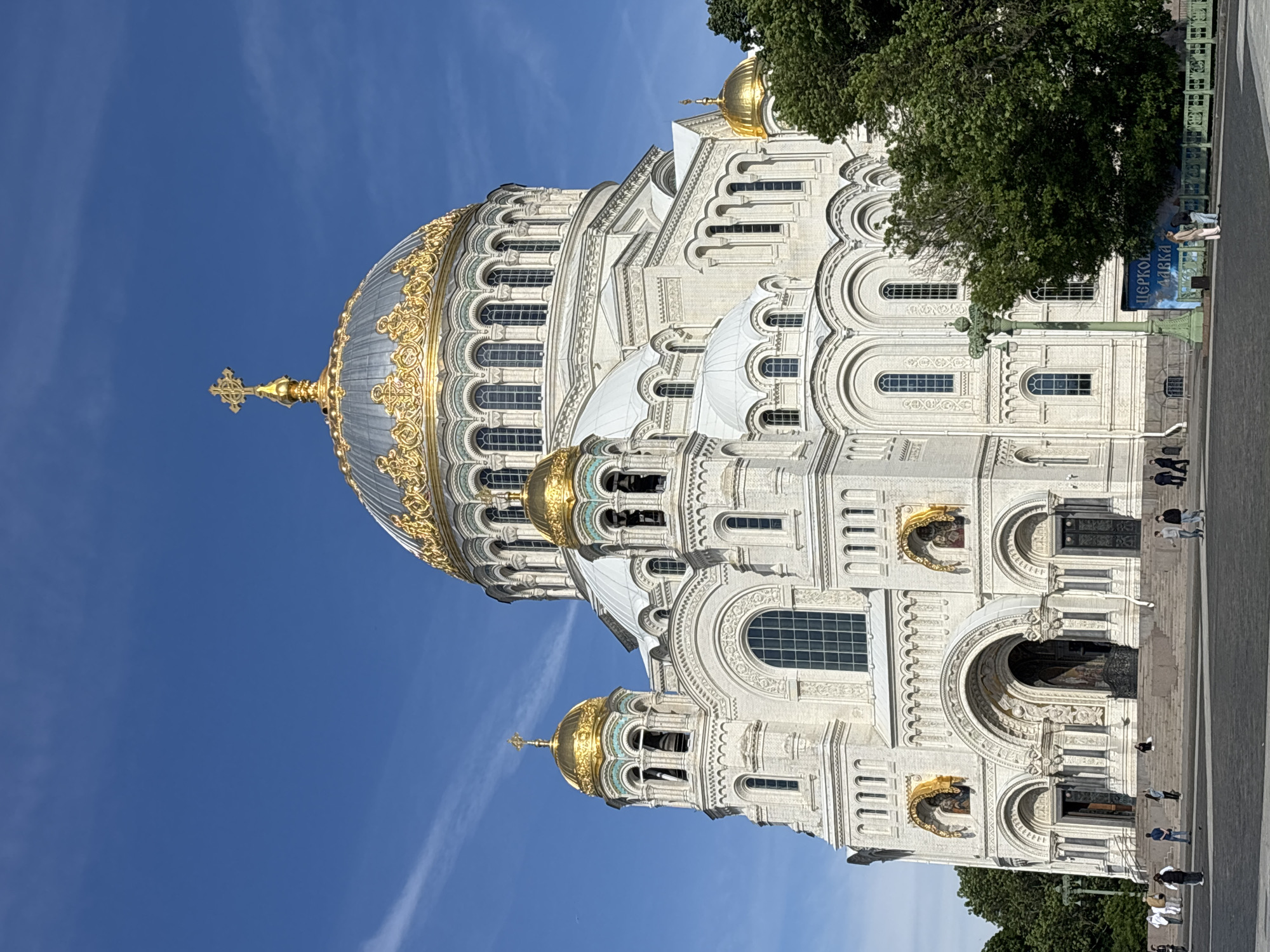 This is an exterior, eye-level, full shot of the Kronstadt Naval Cathedral in Kronstadt, Russia, taken on a clear day under a bright blue sky. The grand, white, brick building features multiple domes and ornate gold accents, most notably the large central dome topped with a golden cross. Smaller golden domes punctuate the structure, adding to its splendor. The cathedral is framed by lush green trees on the right side of the image. In the foreground, a paved plaza is visible, with a few individuals scattered across it. A green lamppost stands to the left of the entrance. The overall scene conveys a sense of architectural grandeur and public space.
