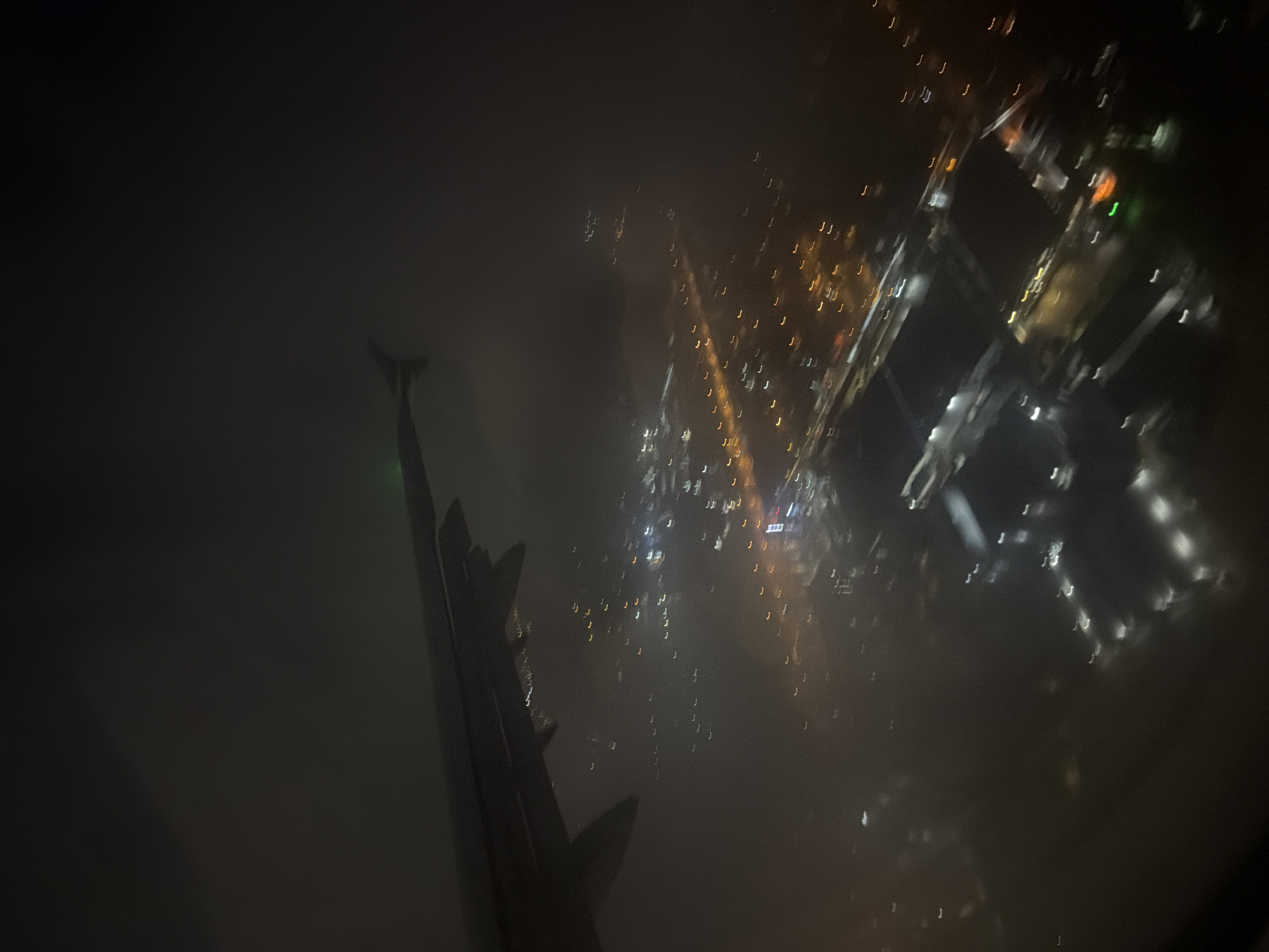 The image is a photograph taken from an airplane window at night, looking down at a city below. The wing of the airplane is visible in the upper left portion of the frame, casting a shadow on the clouds. The city lights are visible as blurred streaks of yellow, white, and orange, indicating movement or a long exposure. Some of the lights appear to be arranged along roads or in clusters, suggesting urban infrastructure. There is a green light visible on the wingtip. The overall impression is one of a city at night, seen through a hazy or cloudy atmosphere from a high vantage point. There are no visible people or specific identifiable landmarks that would pinpoint the exact location within Novoseltsevo, Russia. The scene is dimly lit, with the city lights being the primary source of illumination.