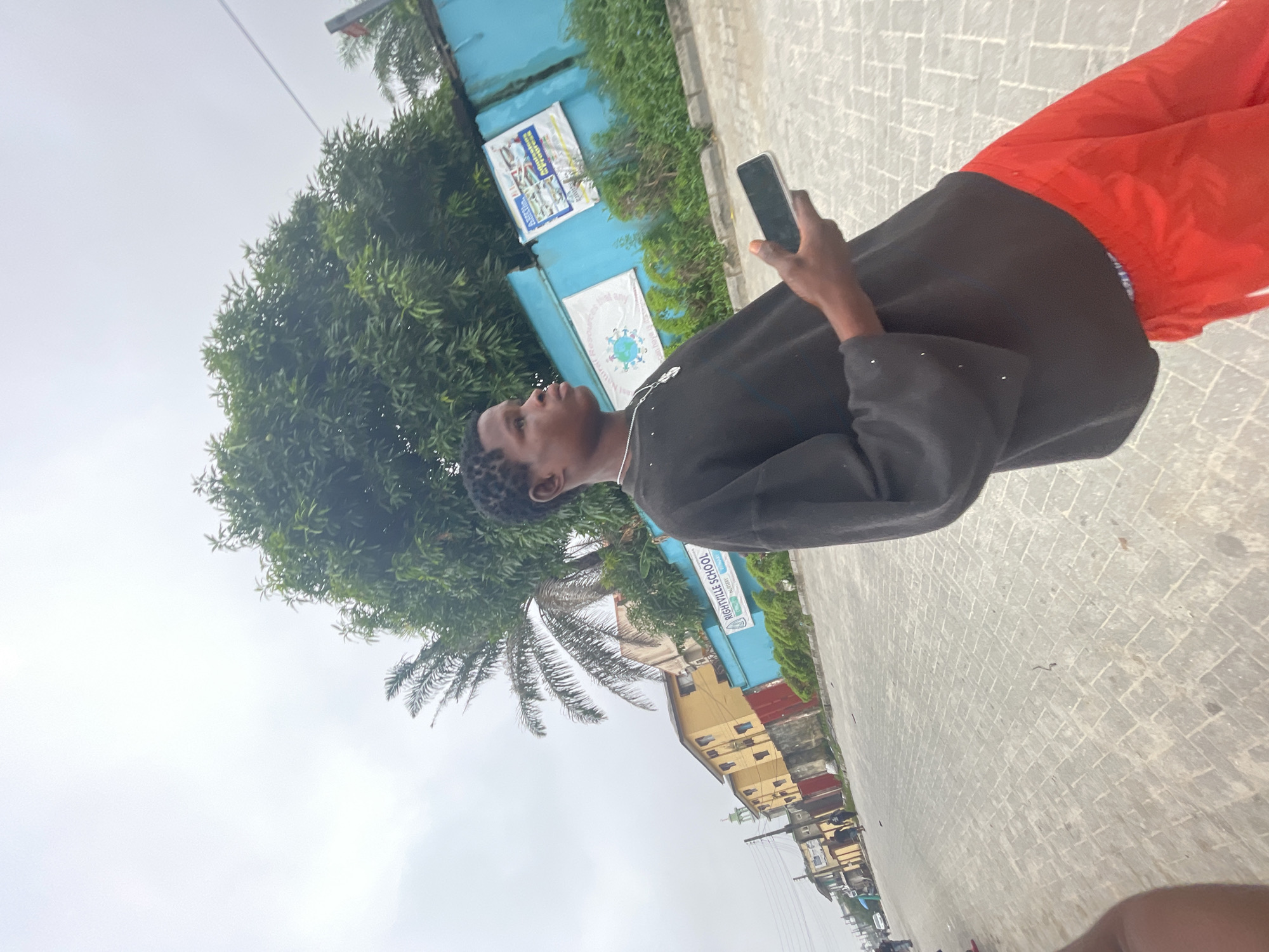 A young male stands on a paved road under an overcast sky in Ado, Nigeria. He wears a dark, loose-fitting, long-sleeved top with small light-colored embellishments on the sleeves, bright red pants, and a silver-colored chain with a circular pendant. He holds a dark-colored smartphone in his right hand and looks towards the right. The road is paved with light gray interlocking blocks. In the background, to the right, a prominent blue wall features multiple signs; one clearly identifies "RIGHTVILLE SCHOOL NURSERY CRECHE." Dense green foliage, including large trees and bushes, is visible behind and above the blue wall. To the left, a series of buildings, including multi-story yellow structures and a lighter building with a green dome, line the street. Utility poles and wires are visible on the left, and distant figures are present on the sidewalk.