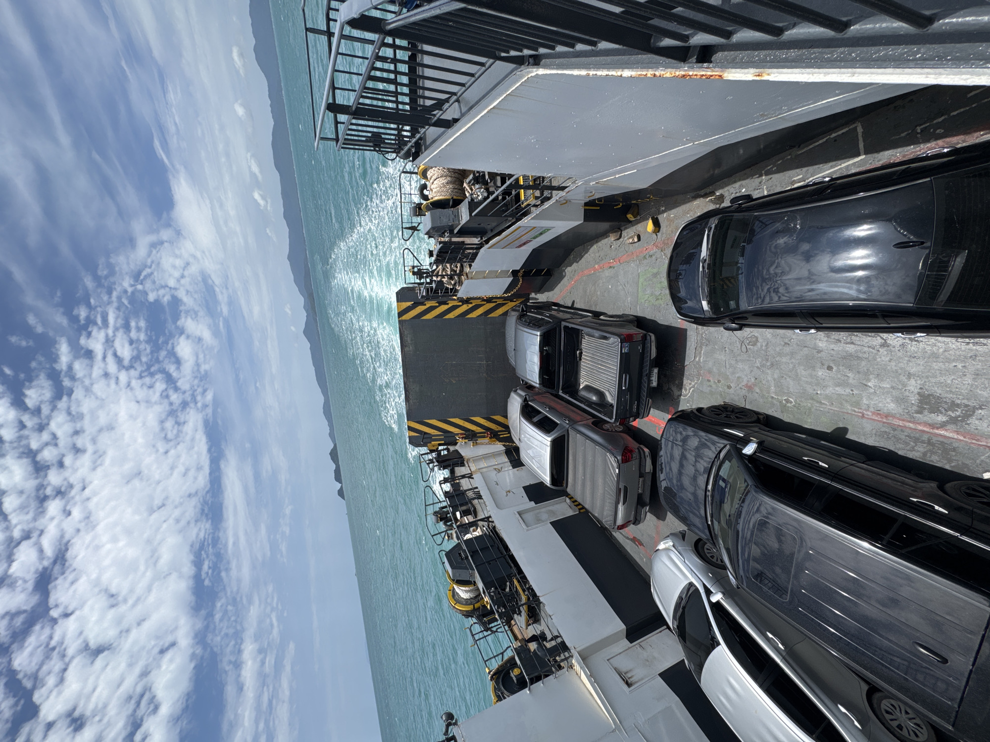 A ferry carrying vehicles moves through turquoise waters under a partly cloudy sky. The ferry deck is lined with several cars, including two pickup trucks, a silver sedan, and two black SUVs. In the background, islands with green vegetation and mountains are visible on the horizon. The ferry is creating a white wake behind it. The image appears to be taken from an elevated position on the ferry, looking forward. The location is specified as Ko Pha-ngan, Thailand.