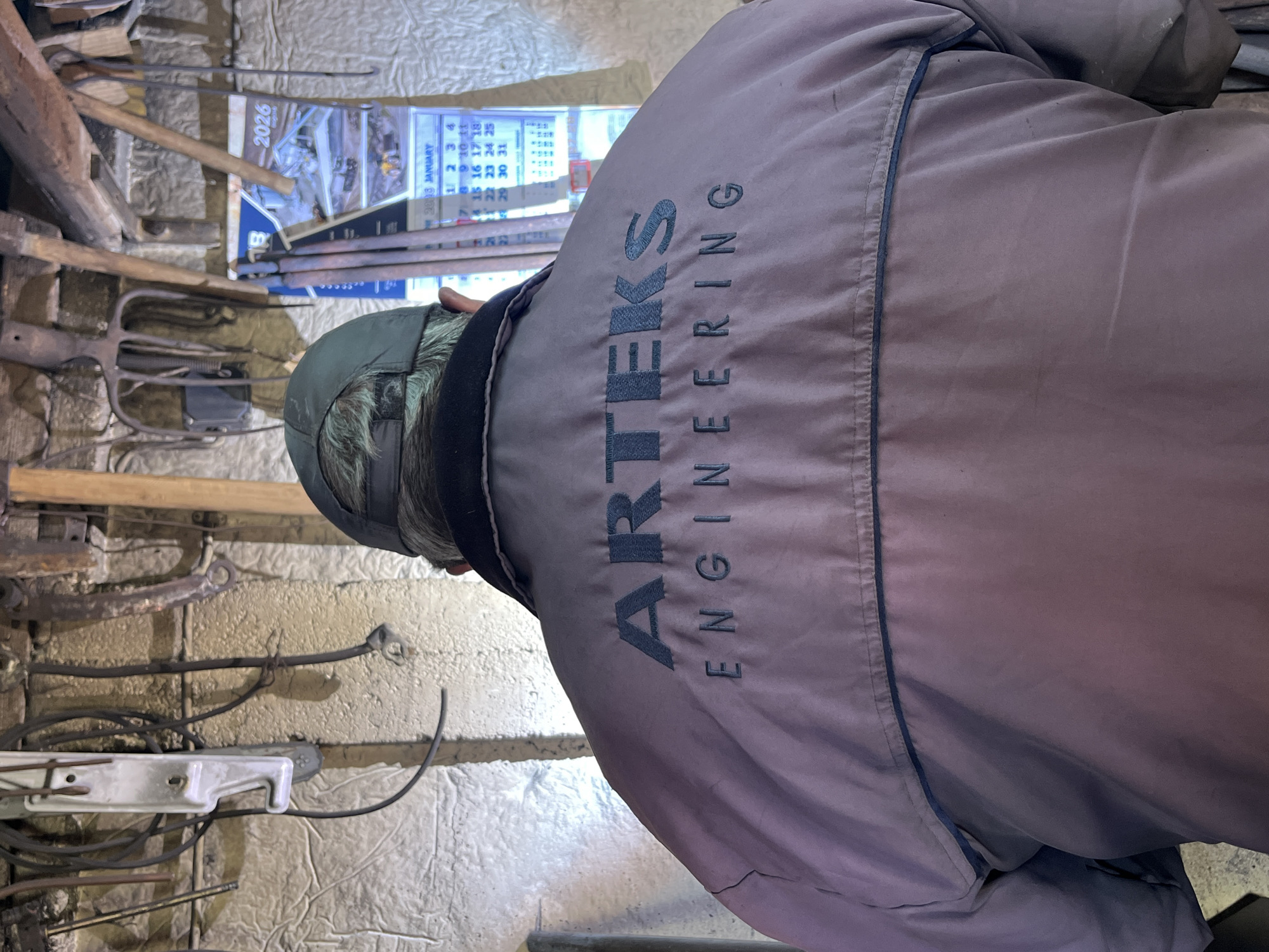 A close-up, slightly angled shot shows the back of a person wearing a grey jacket with the text "ARTEKS ENGINEERING" vertically aligned on the back. The individual has short, grey hair and wears a dark cap. In the background, a cluttered wall is visible with various metal tools and equipment hanging from it. A calendar with the year "2026" and the month "JANUARY" is also visible. The location appears to be a workshop or industrial environment.