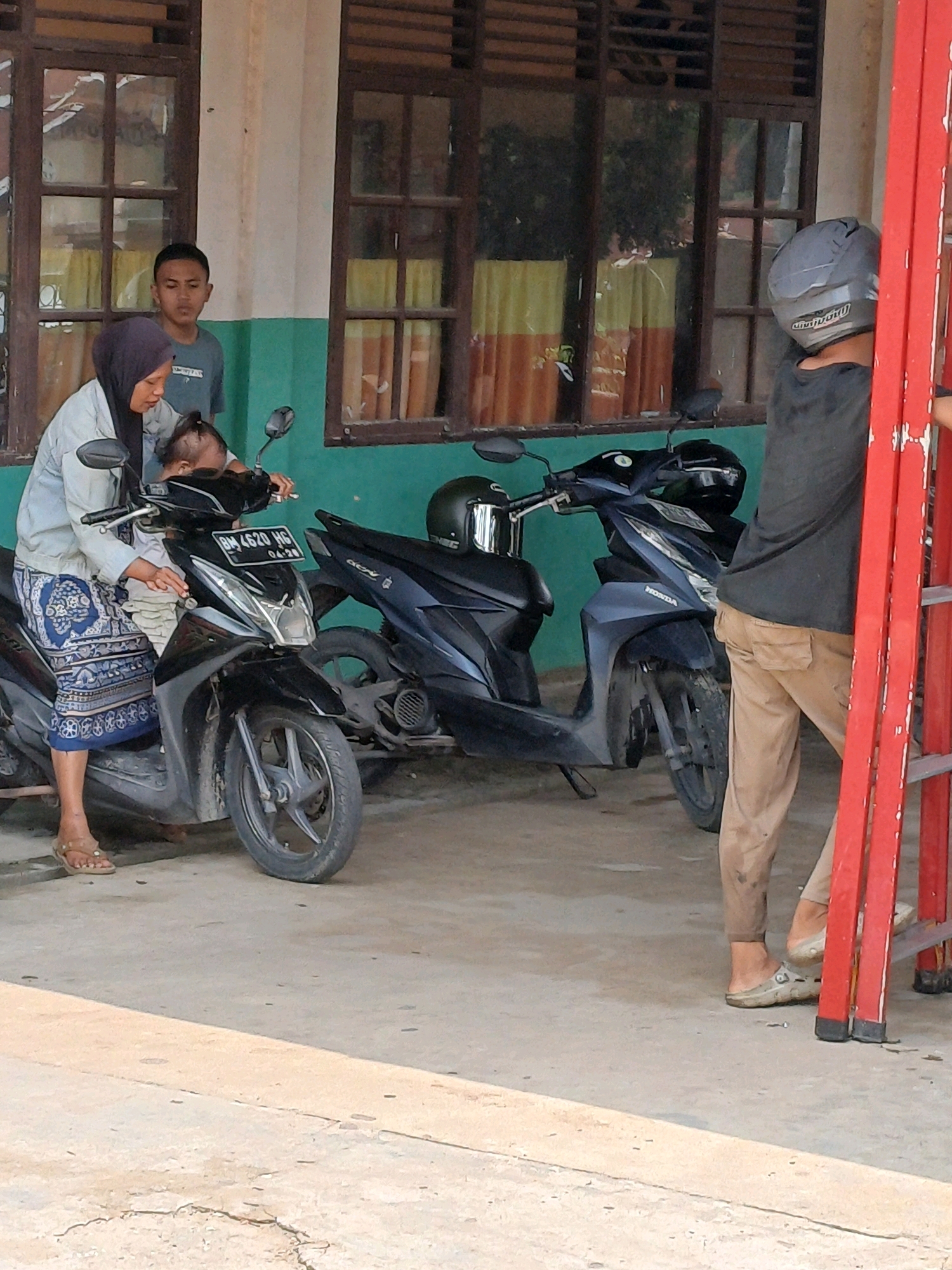 In Dumai City, Indonesia, a group of individuals are gathered near a building with wooden-framed windows. A woman wearing a dark headscarf and a light-colored jacket is seated on a black motorcycle, holding a baby. Another person, a young male wearing a grey t-shirt, stands behind the woman, looking forward. To the right, a person wearing a helmet and a black shirt is standing near a red ladder. Two dark-colored motorcycles are parked side-by-side in front of the building. The ground is paved with concrete.