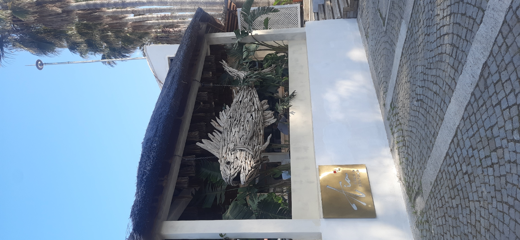 The image depicts the exterior of a restaurant in Kemer, Turkey, as evidenced by the sign featuring Turkish text and the "K" logo. A large wooden fish sculpture hangs from the thatched roof overhang. The building has a white facade. A gold-colored sign is mounted on the wall, displaying stylized script and text indicating "Sushi" and "Seafood." The ground is paved with grey cobblestones. In the background, palm trees are visible against a blue sky.