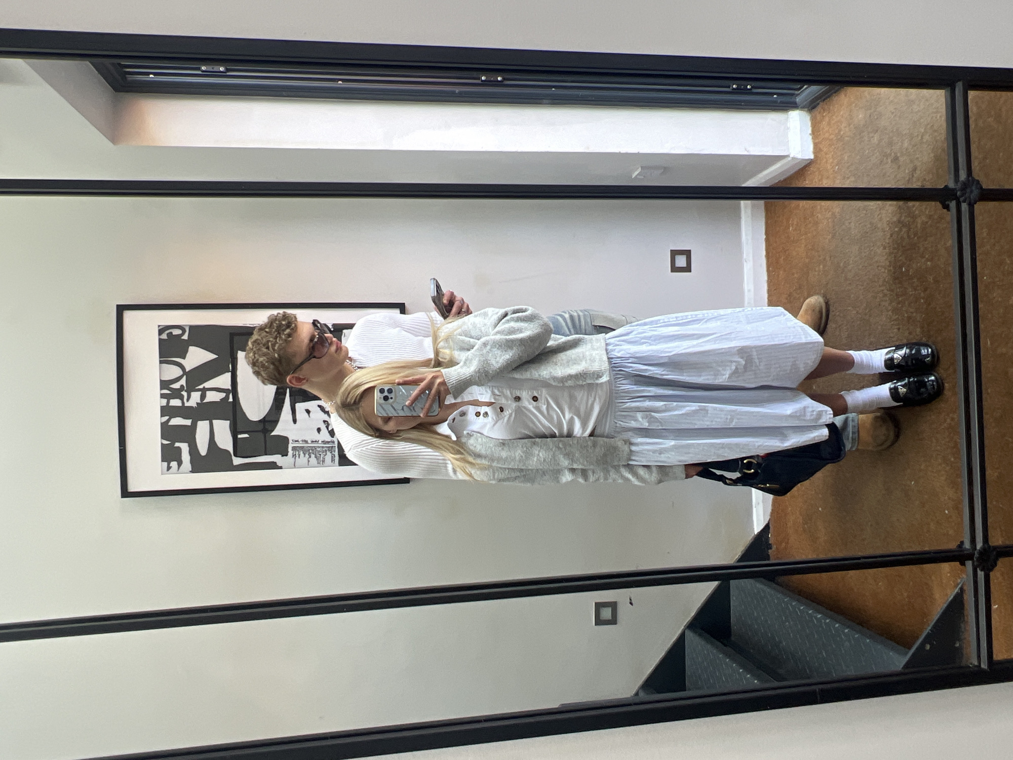 A couple is reflected in a mirror. The man has curly blond hair and sunglasses, and is wearing a white sweater. The woman has long blond hair, and is wearing a striped dress with a gray cardigan. She is taking a photo with her smartphone. In the background there is an abstract black and white painting, a brown floor, and gray metal stairs. The location is possibly in the city of London, United Kingdom.