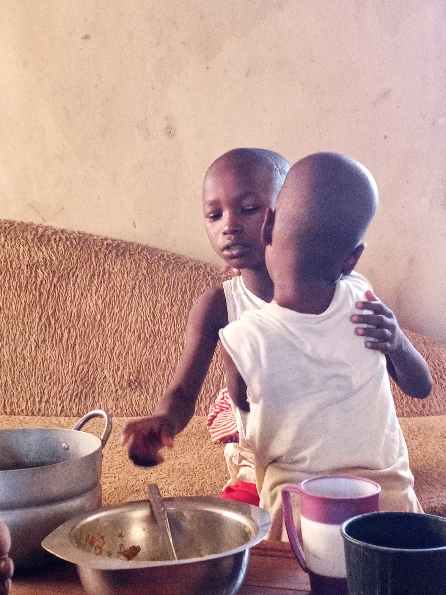 This indoor photograph captures a tender moment between two young children, likely toddlers or preschoolers, with dark skin and shaved heads. They are seated on or in front of a textured, light brown or tan sofa. The background consists of a plain, light-colored wall, which shows some signs of wear and minor blemishes.
In the foreground, several everyday objects are visible on what appears to be a table. To the left, there is a large, metallic pot or bowl with a handle. Centrally placed is a smaller metal bowl containing traces of food and a spoon, suggesting a recent meal. On the right, a purple and white two-toned plastic mug stands next to a dark green or black plastic cup.
The child on the left is partly visible, looking towards the viewer with a neutral expression. Their right hand is extended forward, holding a small, dark, unidentifiable object, possibly a snack. The second child, on the right, is primarily seen from behind, with their head turned towards the first child. This child has their left arm gently wrapped around the first child's waist or back, indicating a close and possibly affectionate interaction. Both children are dressed in simple, light-colored, short-sleeved tops, appearing to be white vests.
The scene conveys a quiet, domestic atmosphere, likely during the daytime due to the natural lighting. The objects and setting suggest a modest home environment in Adonkolo, Nigeria. There is no visible text within the image.