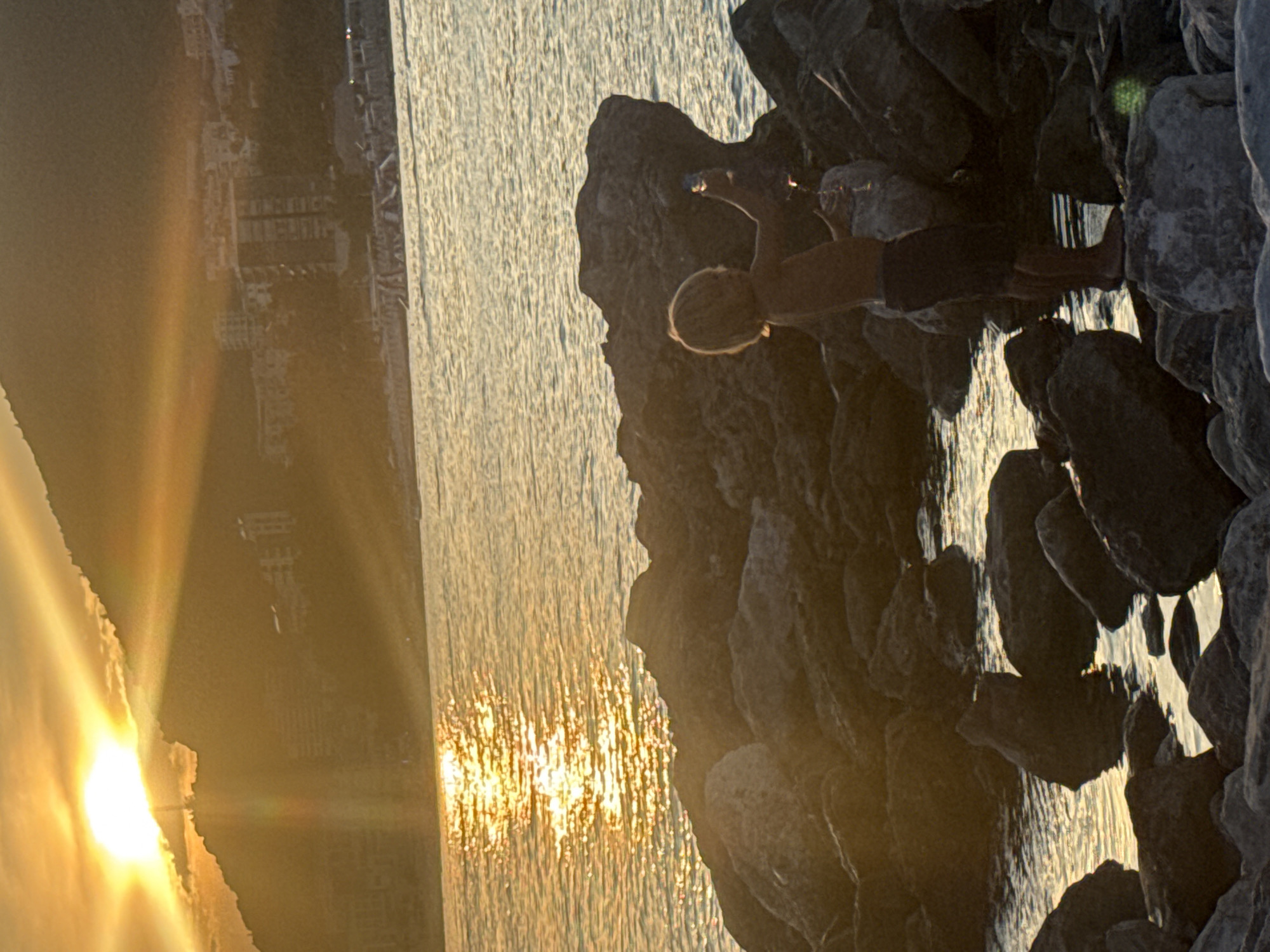 The image depicts a sunset scene along the coast of Rafailovići, Montenegro. A young boy, shirtless and wearing dark shorts, stands on a rocky shoreline, holding a clear plastic bottle and what appears to be a small sparkler. The sun is low in the sky, casting a warm golden glow and creating strong rays that illuminate the scene and reflect off the water. In the background, the town of Rafailovići is visible with buildings nestled along a hillside, partially obscured by the sunset's haze and shadows. The water is calm, with gentle ripples reflecting the sunlight. The foreground is dominated by dark, wet rocks, with the boy positioned on a larger, lighter-colored rock. The overall atmosphere is serene, with the natural beauty of the sunset enhanced by the boy's presence.