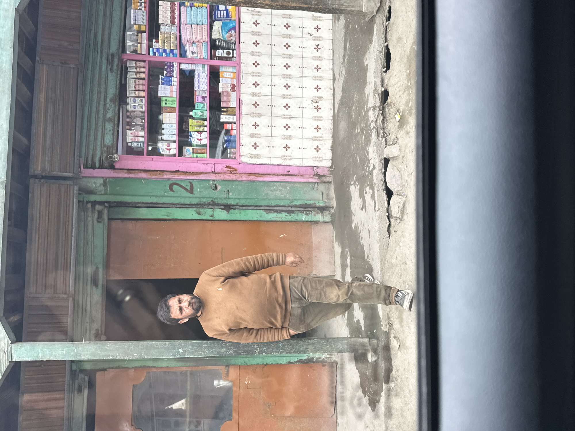 A man walks in front of a shop in Dras, India. The man is wearing a brown sweater and brown pants. He has a beard and is looking to the side. The shop is painted pink and green and has shelves stocked with various products. The ground in front of the shop is wet and muddy. The scene appears to be a street-level view, possibly from inside a vehicle, looking out.