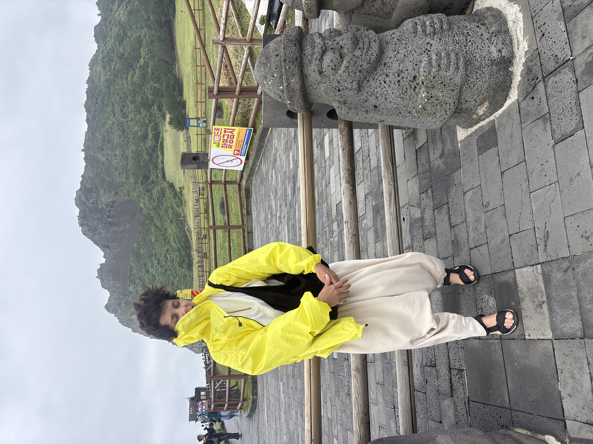 A person wearing a bright yellow raincoat, a light-colored shirt, and loose-fitting cream pants stands in front of a wooden railing. They are also wearing black sandals. The person has their eyes closed and their head tilted down. A black bag is slung over their right shoulder. In the background, a grassy hillside with a rocky peak rises under a cloudy sky. A wooden fence runs across the mid-ground. To the right of the person, a grey, rough-hewn stone statue with a hat and carved facial features stands beside the railing. Another similar statue is partially visible to the right. A sign with Korean text and a red circle with a diagonal line is posted on the fence. The ground is paved with dark grey interlocking stones. This scene is located at Seongsan Ilchulbong Peak in Seogwipo-si, South Korea.