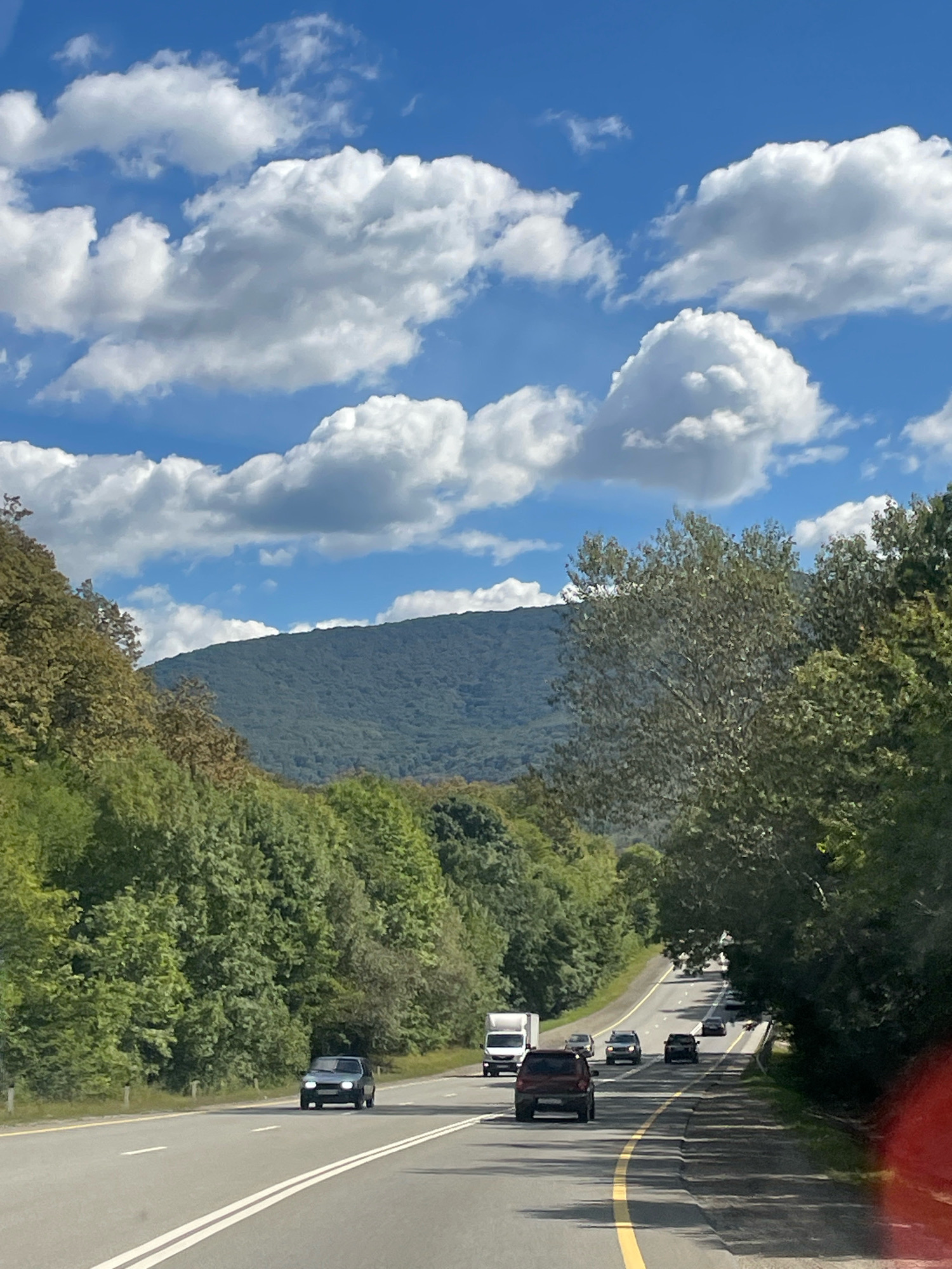This is a photograph taken from a moving vehicle on a highway in Defanovka, Russia. The highway curves to the right, bordered by lush green trees on both sides. In the distance, a large, tree-covered mountain range dominates the landscape under a bright blue sky filled with fluffy white clouds. Several vehicles are visible on the road, including cars, a white truck, and a smaller white van. The road has clear white lane markings and a yellow line marking the edge. The sun is shining, casting shadows from the trees onto the road, suggesting it is daytime.