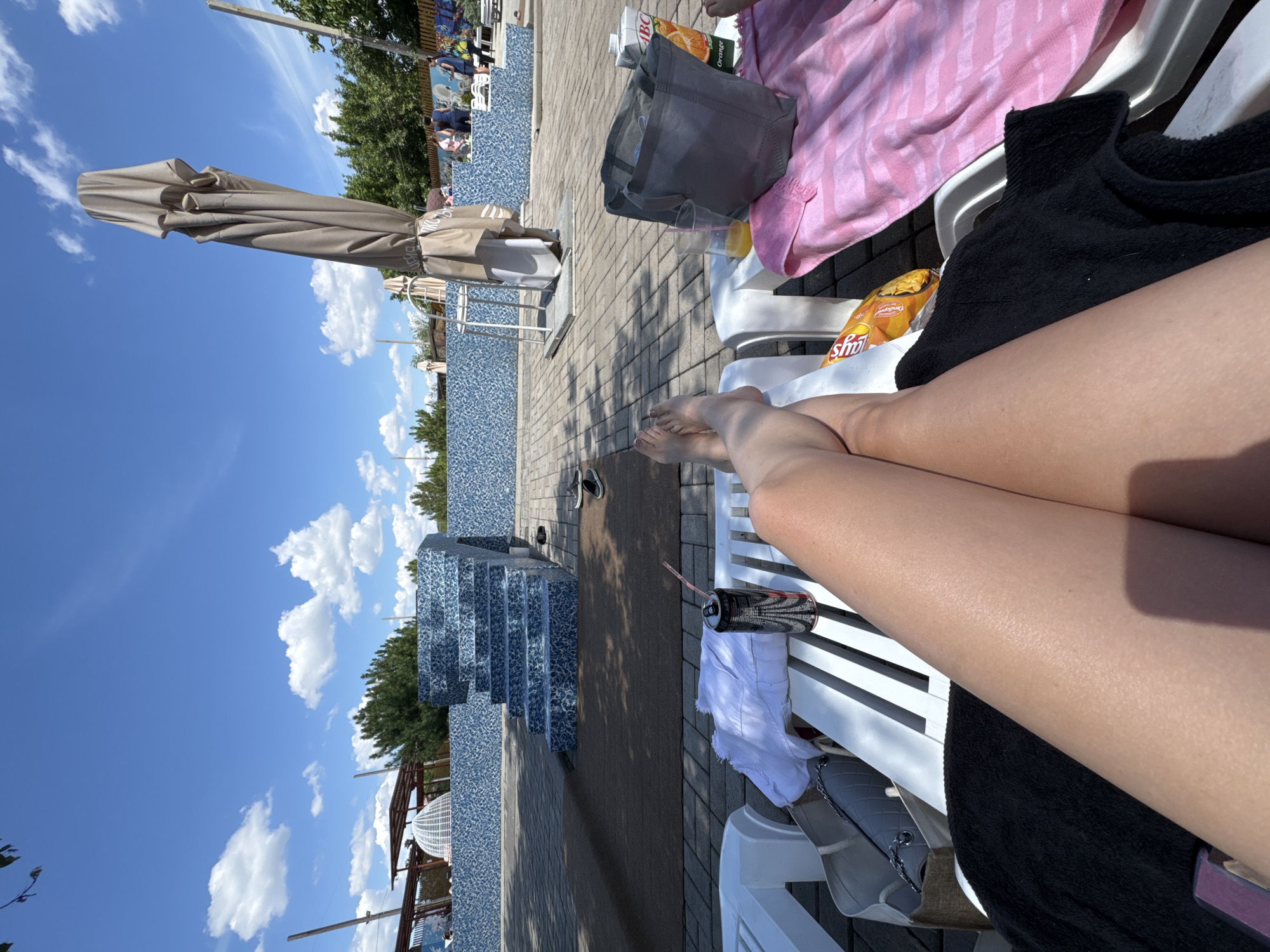 The image depicts a sunny day at an outdoor swimming pool area, possibly a resort or recreational facility. In the foreground, a person's legs are extended across two white lounge chairs. One chair has a black towel draped over it, and the other has a pink striped towel, a gray tote bag, a carton of orange juice, a glass with a straw, and a bag of chips. A can with a straw and a white towel are on the same chair as the legs. In the background, there are blue tiled steps leading into a pool, a closed umbrella, and some trees. Several people can be seen in the middle ground, some standing and some sitting on lounge chairs. The sky is clear blue with scattered white clouds. The location is identified as Makiivka, Ukraine.