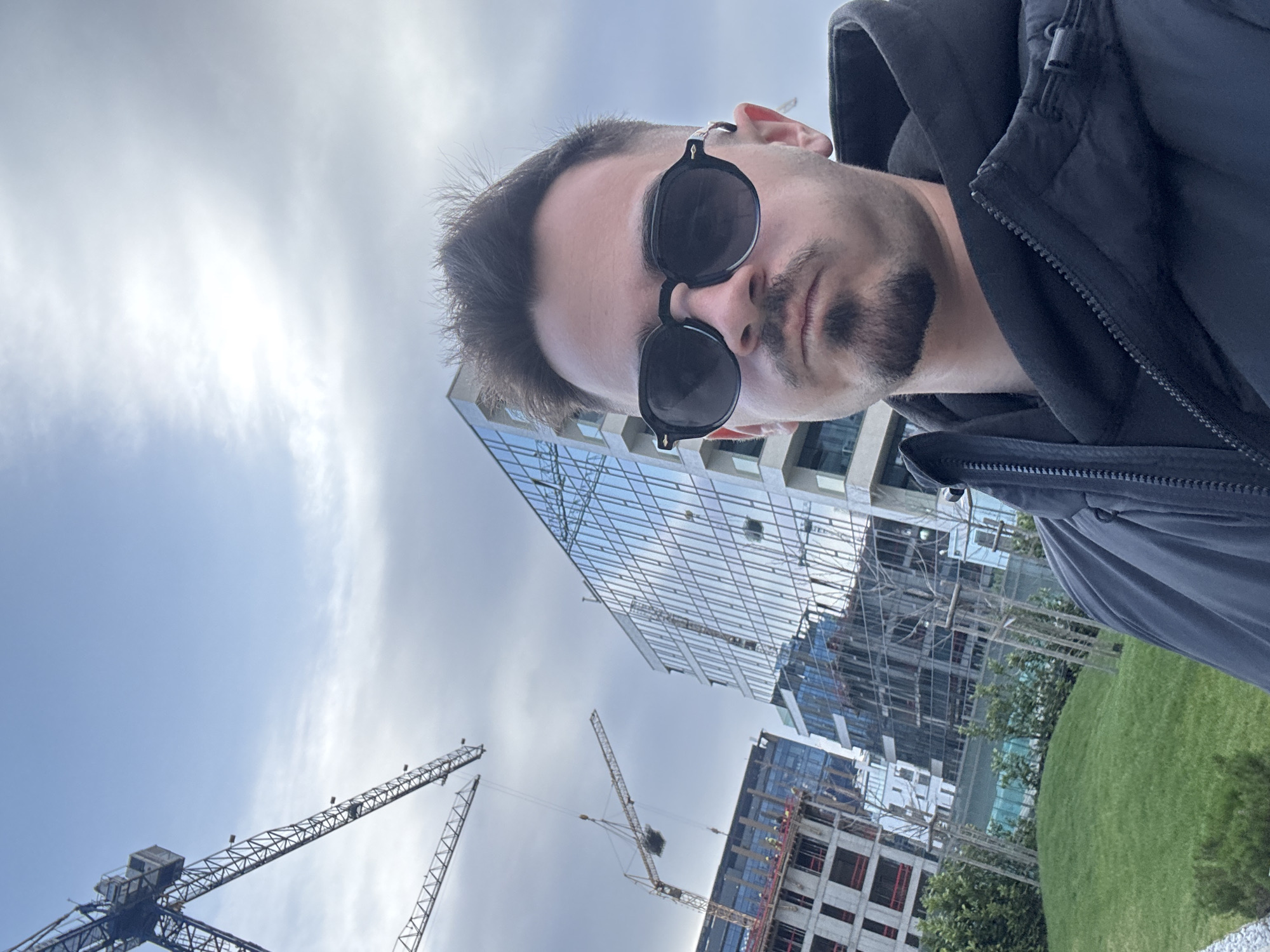 A man with dark hair, a goatee, and sunglasses is facing the camera in an outdoor setting. He wears a dark jacket. Behind him, there are several modern buildings, some of which appear to be under construction, as indicated by the presence of construction cranes. There is also a patch of green grass visible. The sky is overcast. It's hard to tell for sure without clear landmarks, but based on the modern architecture and construction activity, the location could possibly be in a newer developing area of Sofia, Bulgaria, such as around Business Park Sofia.