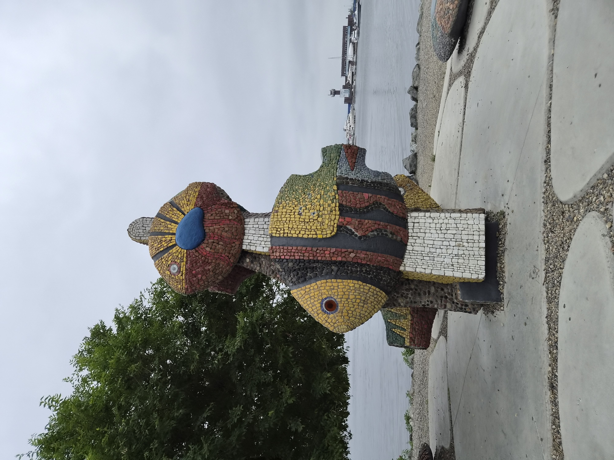 This is a medium-shot photograph of a colorful mosaic sculpture of a fish-like creature situated in an outdoor public space. The sculpture is made of many small, irregular tiles in shades of yellow, red, blue, black, and white, creating a vibrant, abstract design. The creature has a large, round blue eye with a red pupil and a yellow-patterned body. It is positioned on a concrete platform with a decorative border of small pebbles.
In the background, a large green tree with dense foliage is visible on the left side of the frame, partially obscuring the sky. Beyond the tree and sculpture, a body of water, likely the sea, stretches to the horizon. On the far side of the water, a port or harbor area is visible with several white yachts and boats docked. A tall, modern tower with a lighthouse-like structure at its top stands prominently in the harbor, along with a long, low building. The sky is overcast and gray, suggesting a cloudy day. The overall scene conveys a sense of a public waterfront park or promenade.
This sculpture is located at the "Malchishnik" (Bachelors') embankment in Vladivostok, Russia. It is part of the seaside promenade's artistic design.